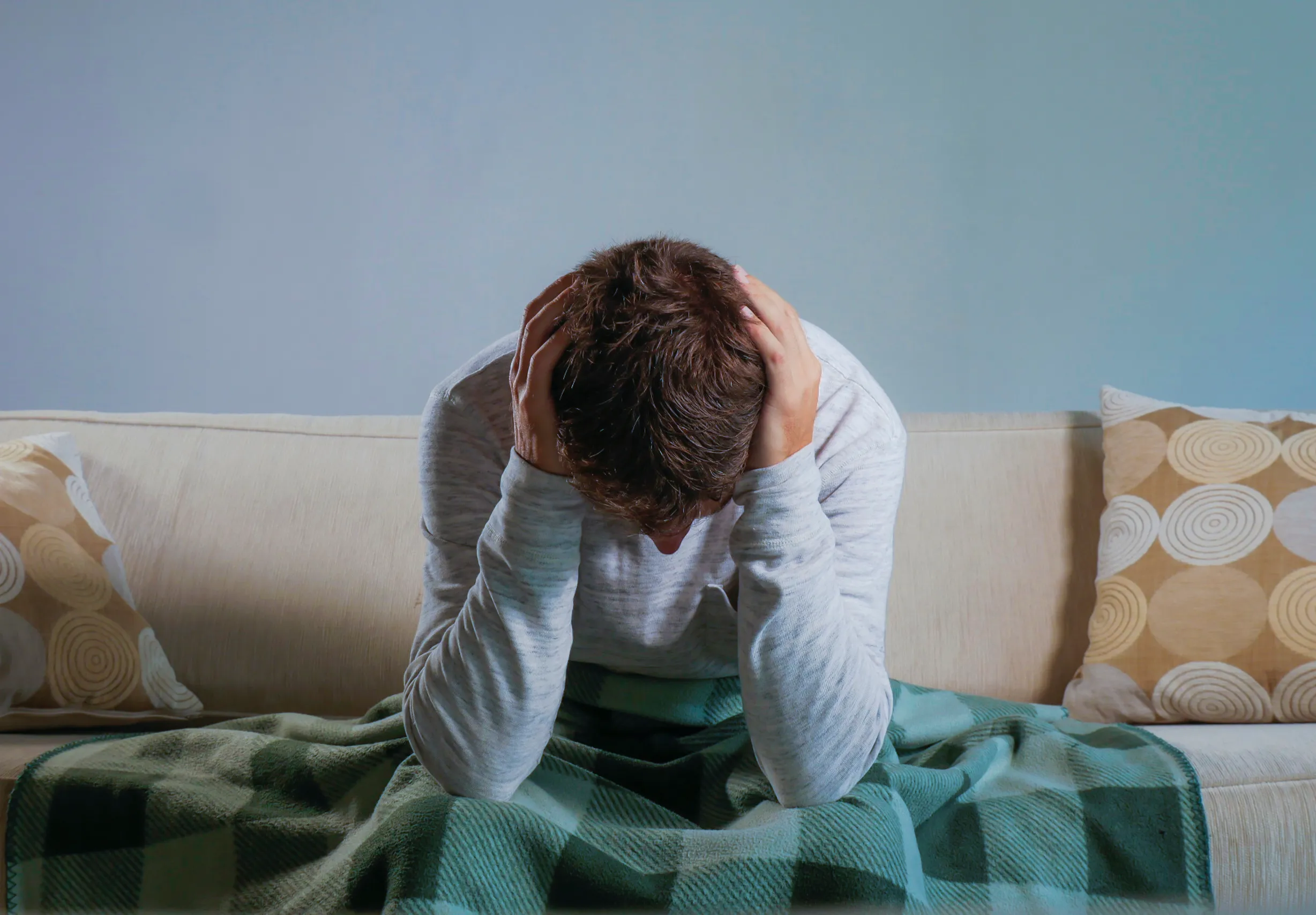 A person sits on a couch with their head in their hands, appearing upset and perhaps struggling with anxiety. They are covered with a green plaid blanket. Two cushions with circular patterns are on either side of the couch, contrasting against the light blue background wall.