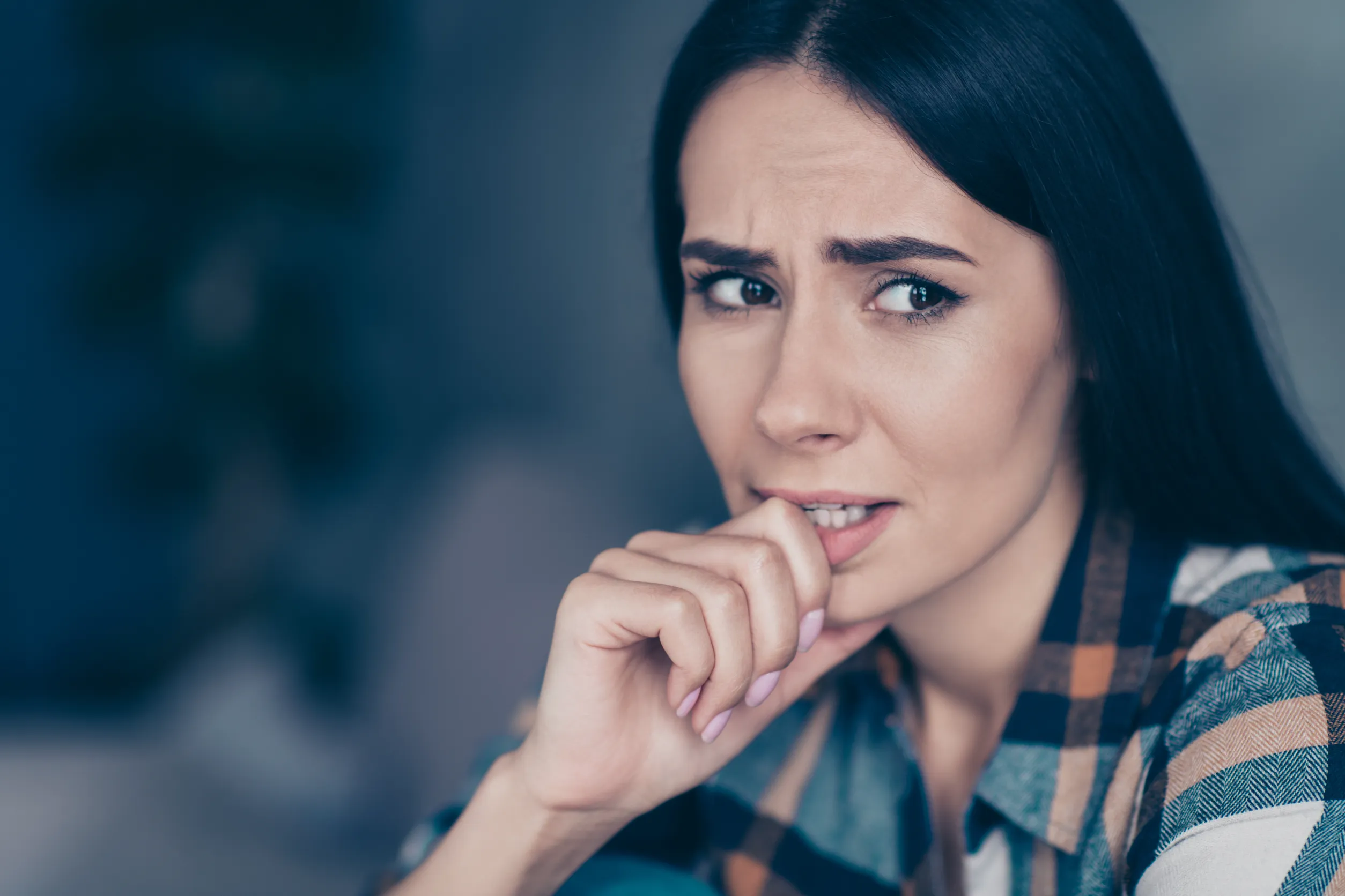 A woman with long dark hair and a checkered shirt appears anxious, biting her nails while looking off to the side. The background is blurred, emphasizing her worried expression, highlighting the importance of understanding mental health.