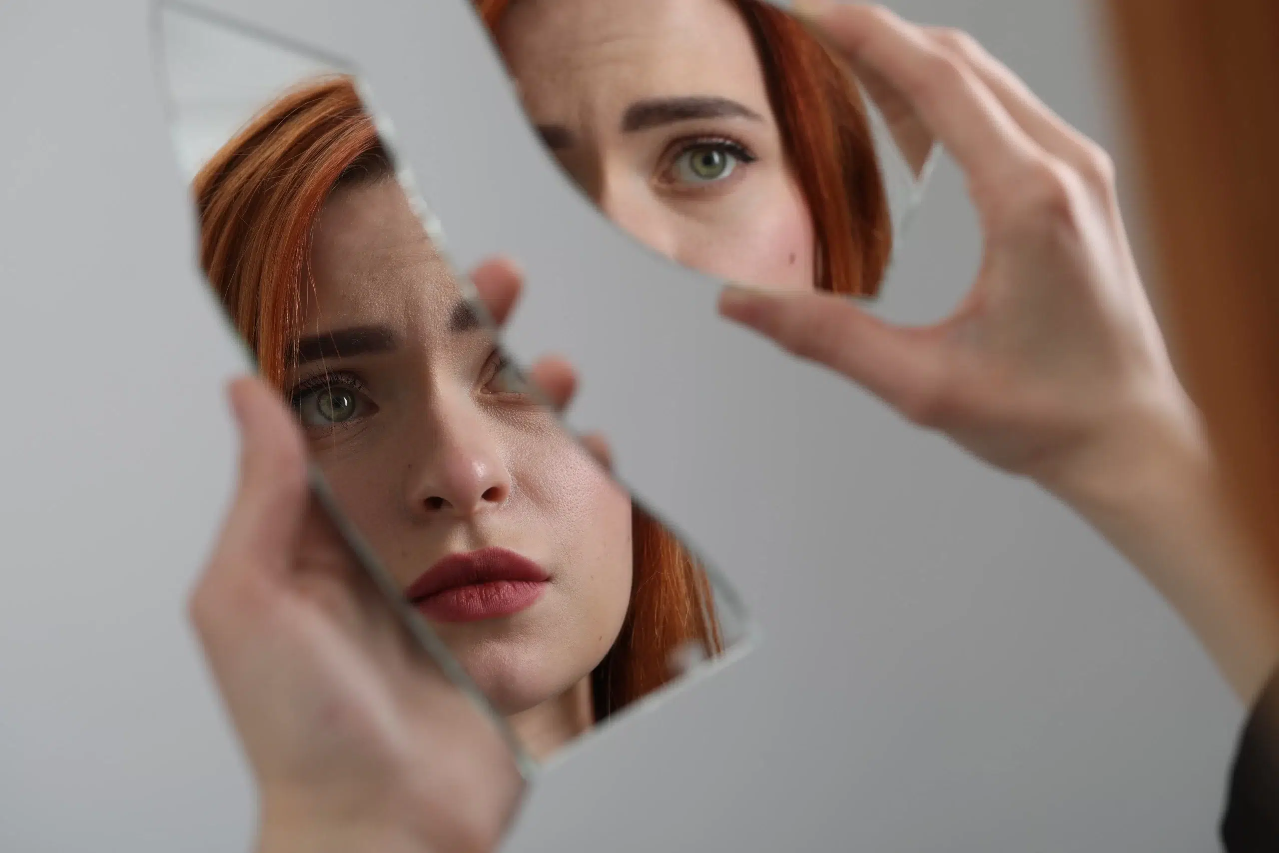 A person with red hair looks intently at their reflection in a broken mirror, holding a piece carefully. The background is neutral, emphasizing the reflective shards and the person’s contemplative expression, subtly hinting at signs often associated with bipolar disorder.