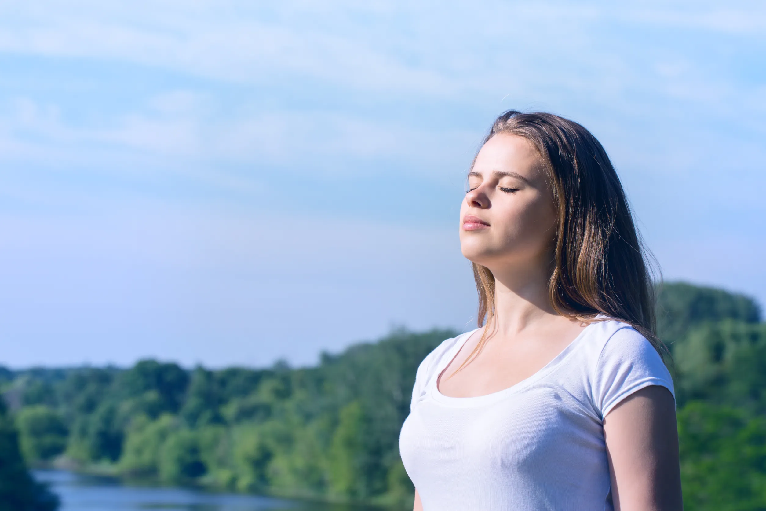 A woman with long brown hair stands outdoors with her eyes closed, managing stressors by soaking in the sun. She is wearing a white t-shirt, enjoying the bright, clear day. Trees and a river are in the background under a blue sky.