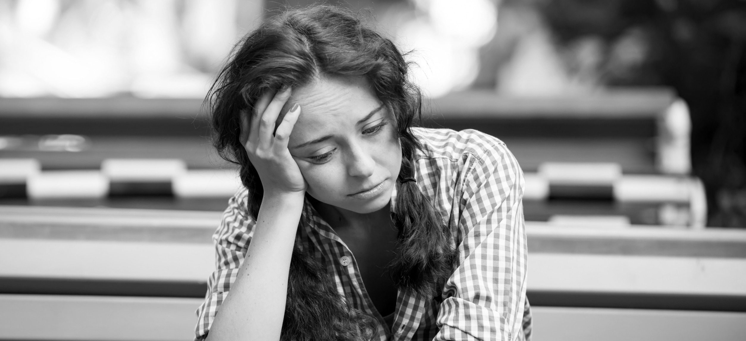 A woman with long hair, clad in a checkered shirt, sits at a table, her head resting on her hand as she gazes down contemplatively, caught in auto draft of thoughts. The blurred background hints at an outdoor setting, and the black and white hues capture the depth of her mood.