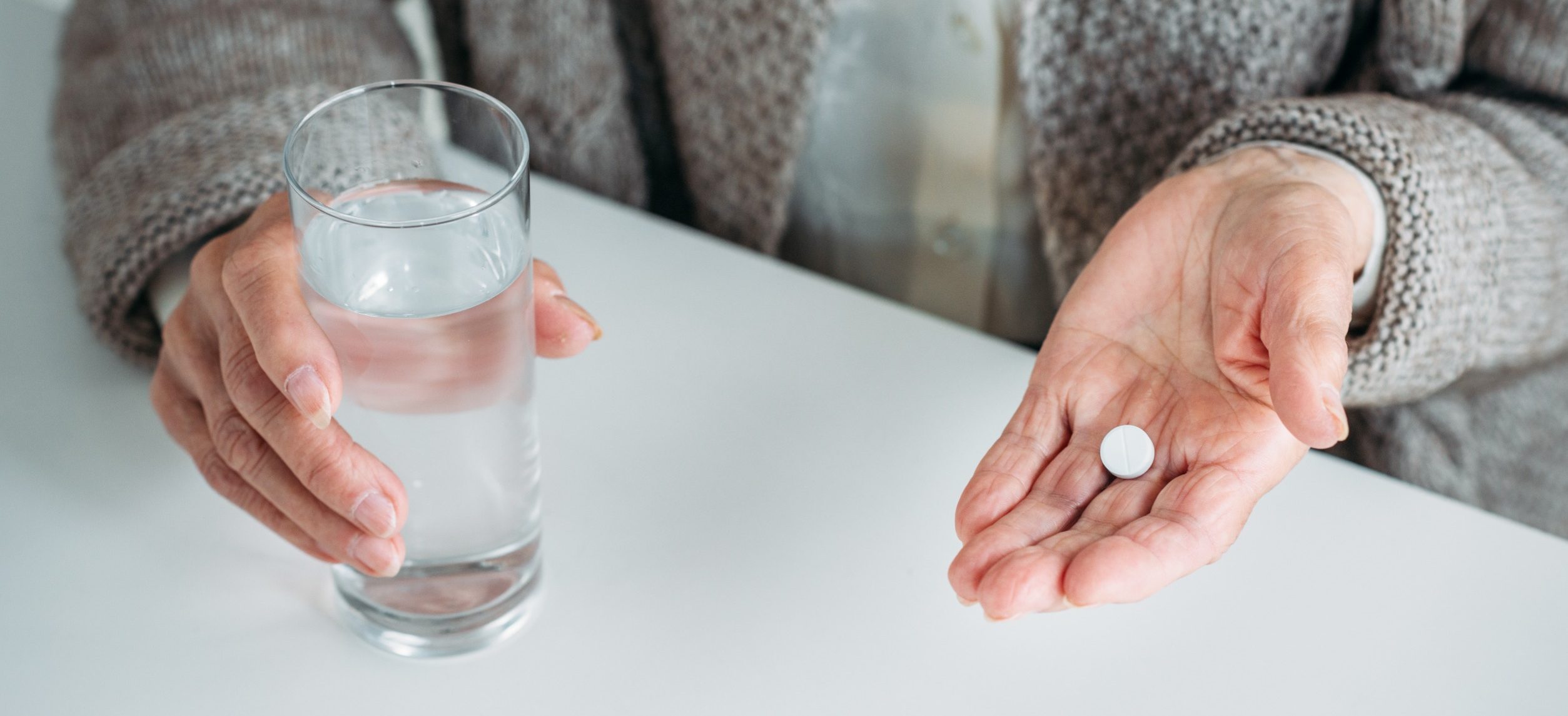 A person holding a glass of water in their left hand and a white pill in their right hand, dressed in a cozy sweater, sitting at a white table.