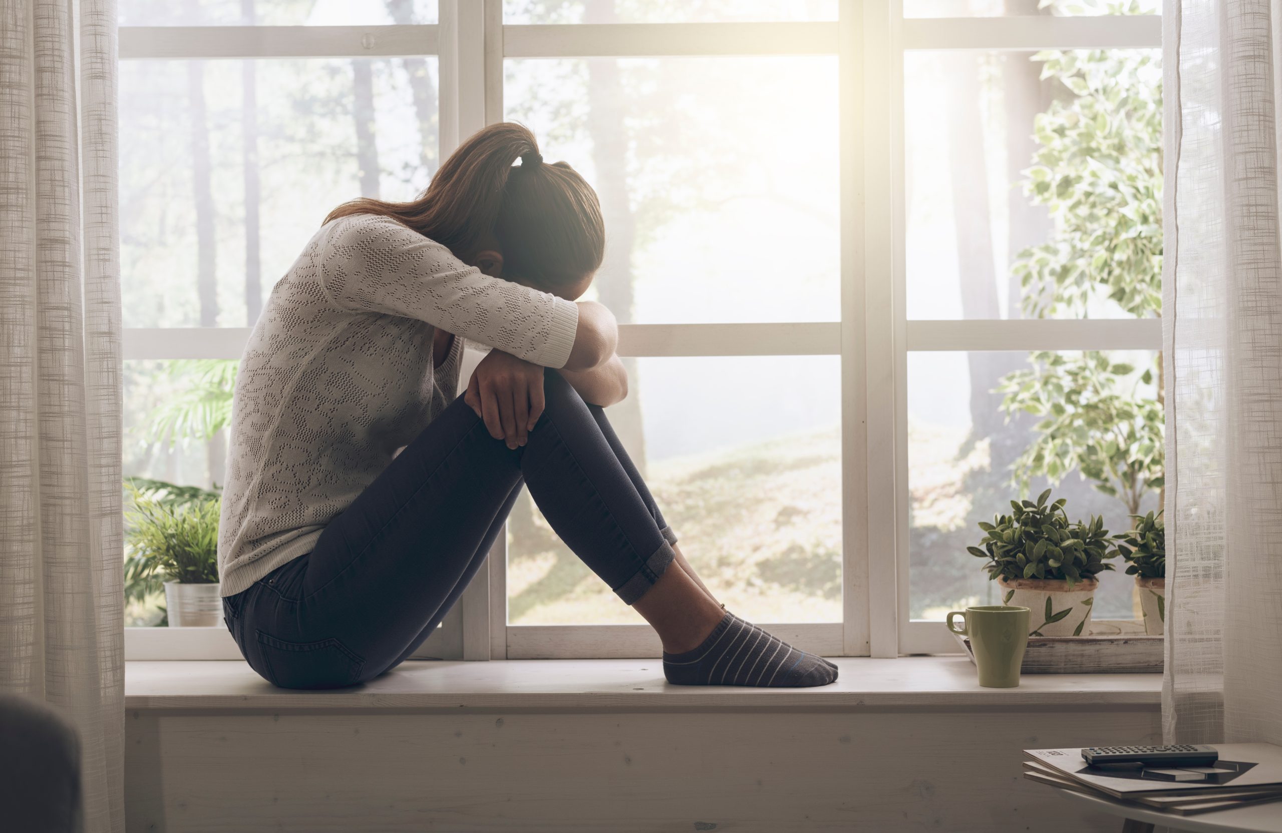 A woman sits on a windowsill with her head resting on her arms, looking pensive. She wears a white sweater, jeans, and socks. Sunlight filters through the window, casting a gentle glow over the room. Potted plants and a mug sit nearby.