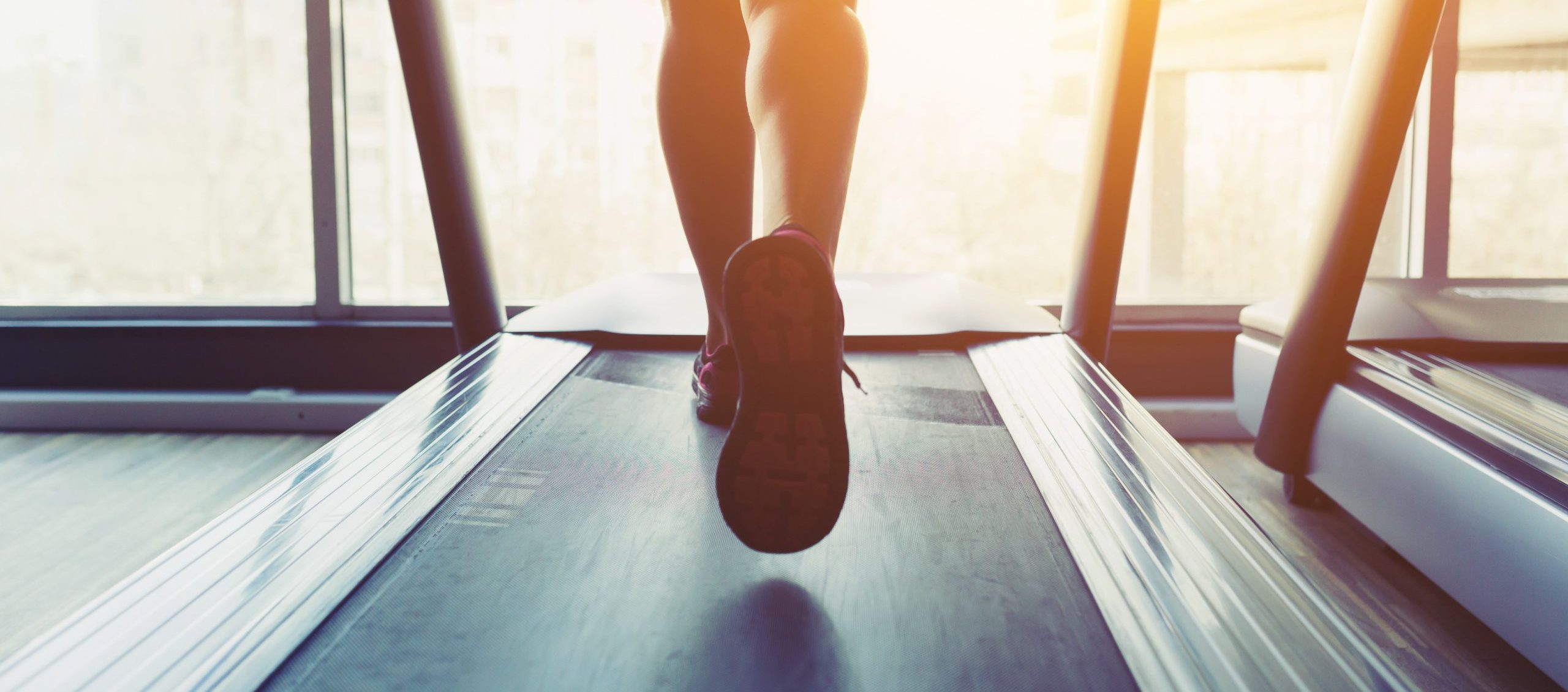 A person runs on a treadmill indoors, embracing the connection between physical health and mental health, with bright sunlight streaming through large windows in the background. Focus is placed on their lower legs and the treadmill surface.