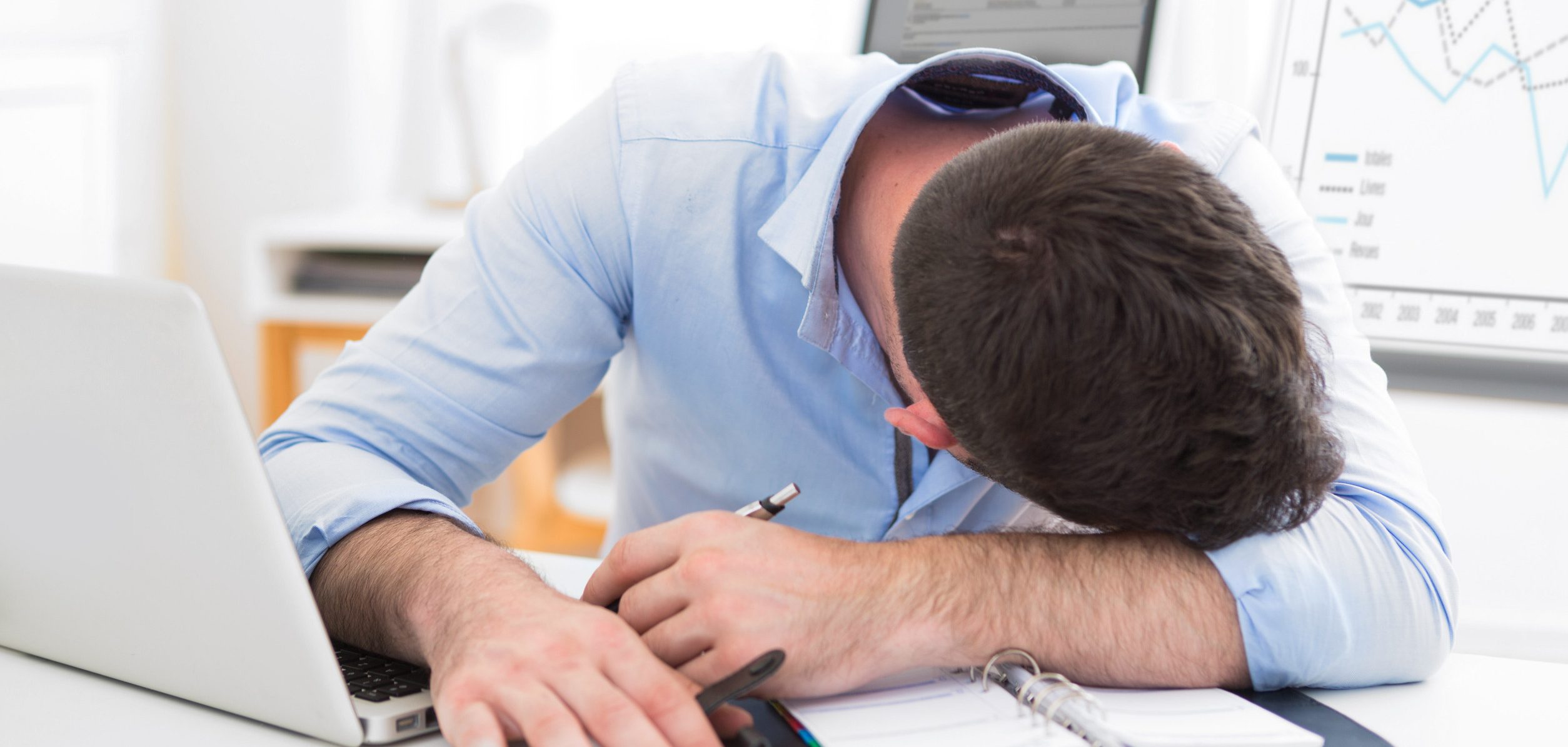 A man in a blue shirt rests his head on a desk with a laptop, glasses, and documents. A graph on a screen in the background shows fluctuating lines, indicating data analysis or work.