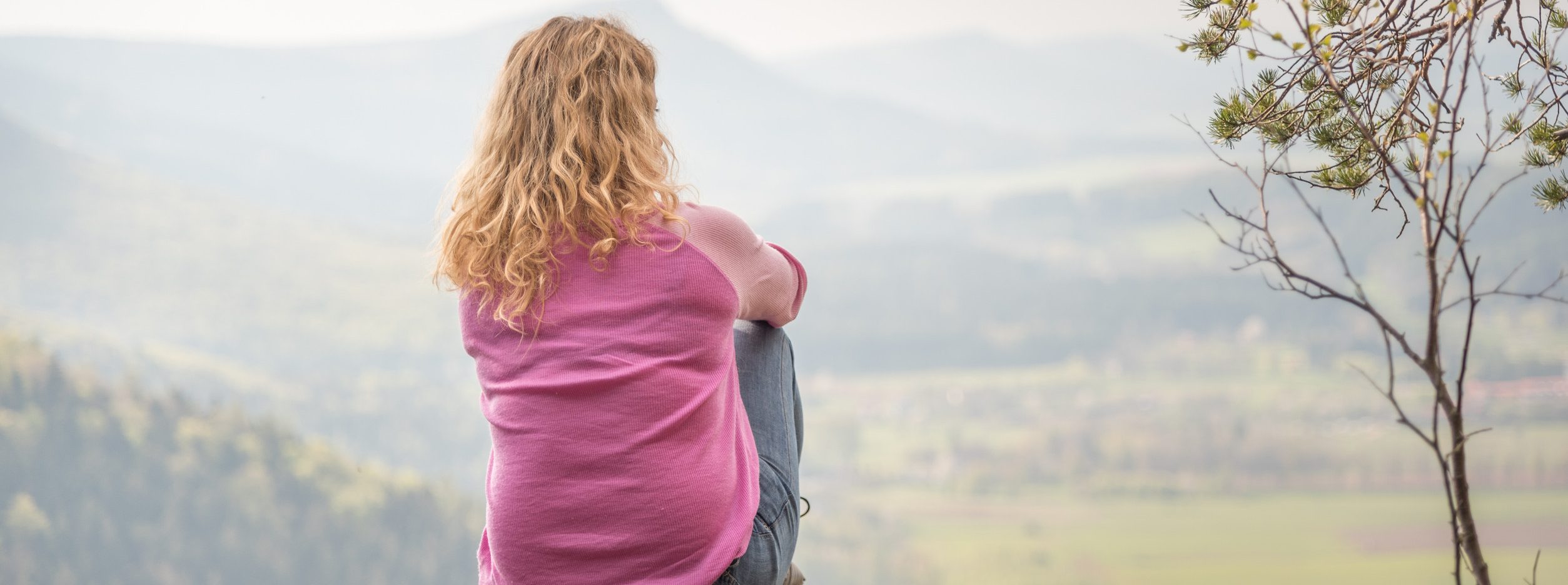 A person with long curly hair, wearing a pink top and jeans, sits mindfully on a rock overlooking a vast landscape of rolling hills and greenery under a cloudy sky. A tree with sparse leaves is visible to the right, reflecting the balance found in nature.