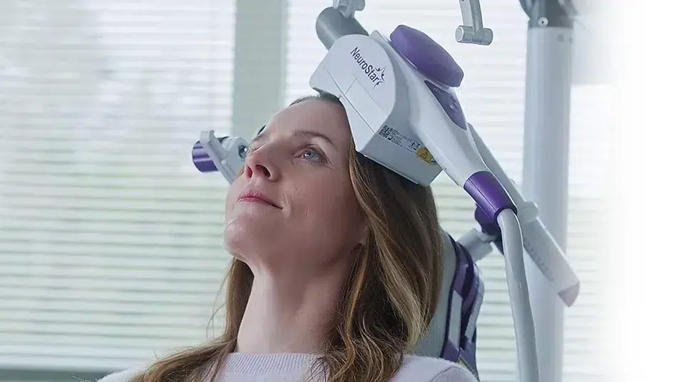 A person sits calmly with a medical device positioned around their head, suggesting TMS Therapy related to neural activity. They are seated in a bright room with window blinds in the background, possibly seeking long-term benefits for their mental health.