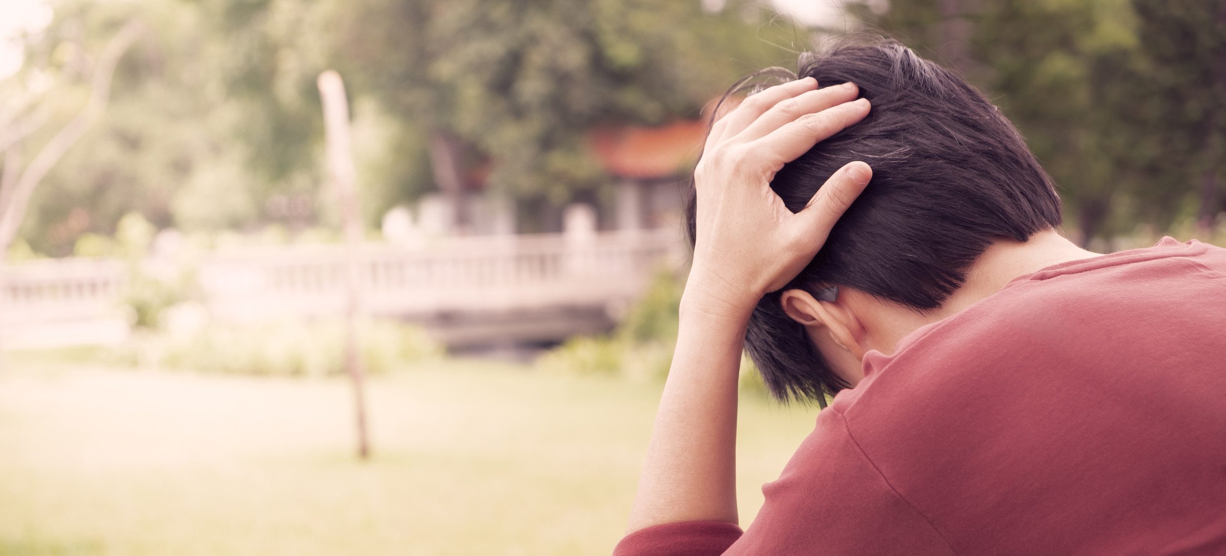 A person sitting on grass with their head in their hands, wearing a red shirt. The background is blurred and shows trees and structures, suggesting a park setting.