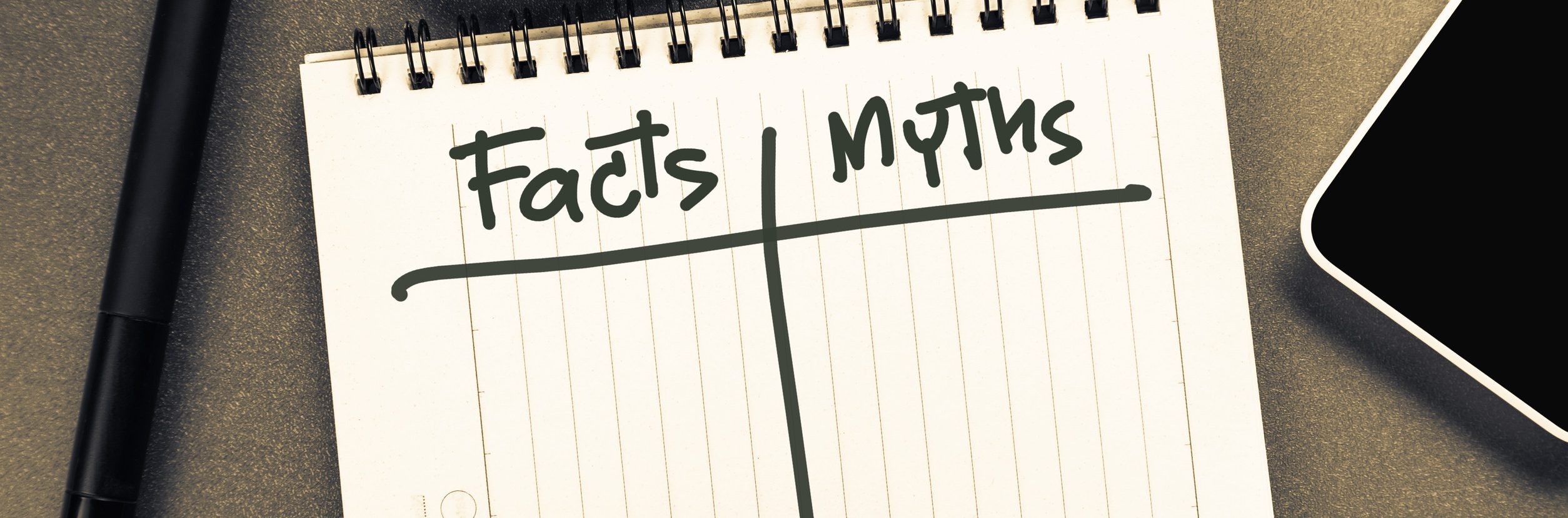 A notebook with two columns labeled "Facts" and "Myths" is placed on a table. A pen lies to its left, and the corner of a smartphone and a tablet are partially visible. The scene is in sepia tones.