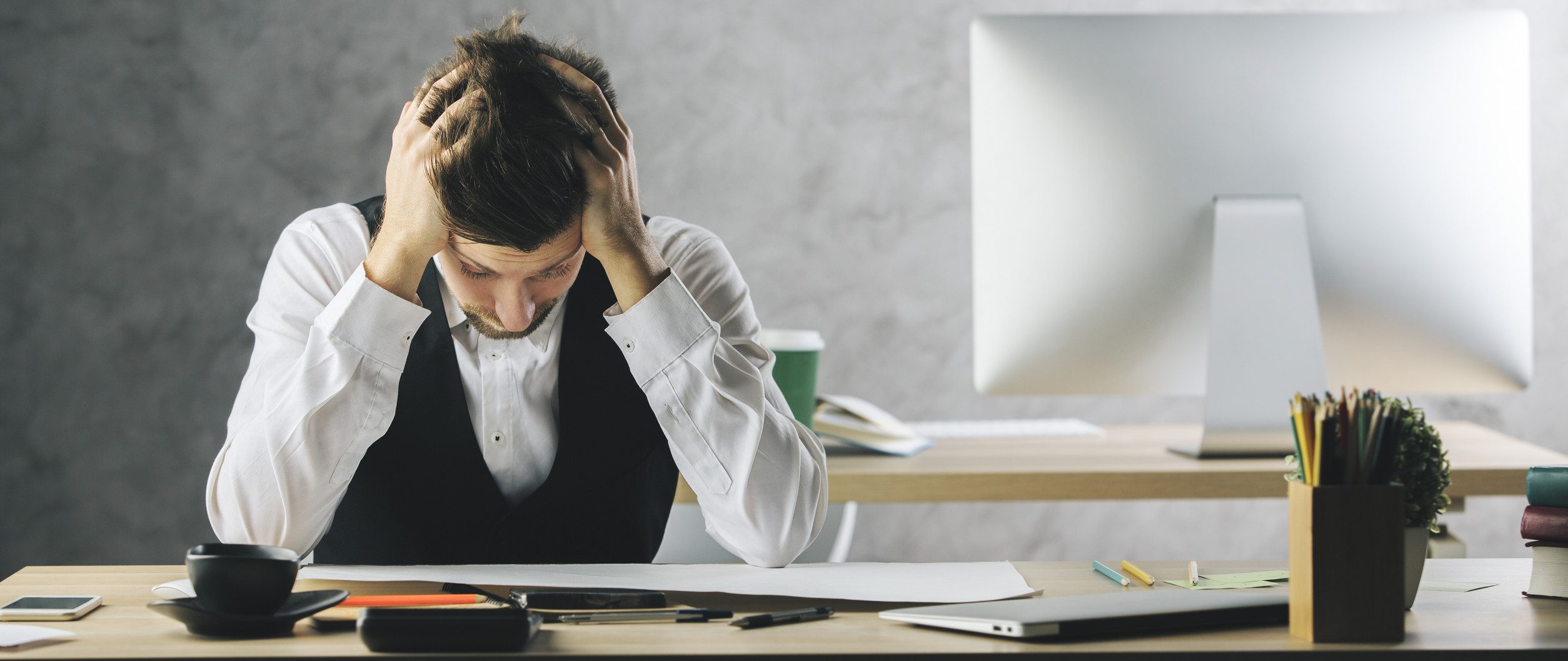 A man, grappling with work stress, sits at a desk with his head in his hands. He is dressed in a white shirt and vest. On the desk are a computer, a cup, stationery, and folders—a stark reminder of the mental health challenges many face against the backdrop of a plain gray wall.