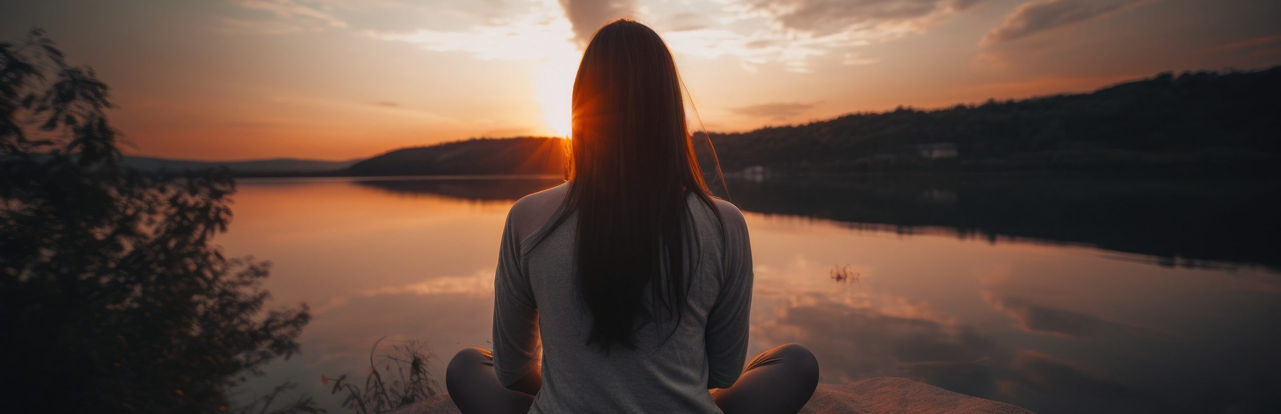 A person sits cross-legged on a blanket facing a tranquil lake, prioritizing self-care by watching a vibrant sunset. The sky is filled with colorful clouds reflecting on the water, creating a peaceful and serene atmosphere. Silhouettes of trees line the horizon.