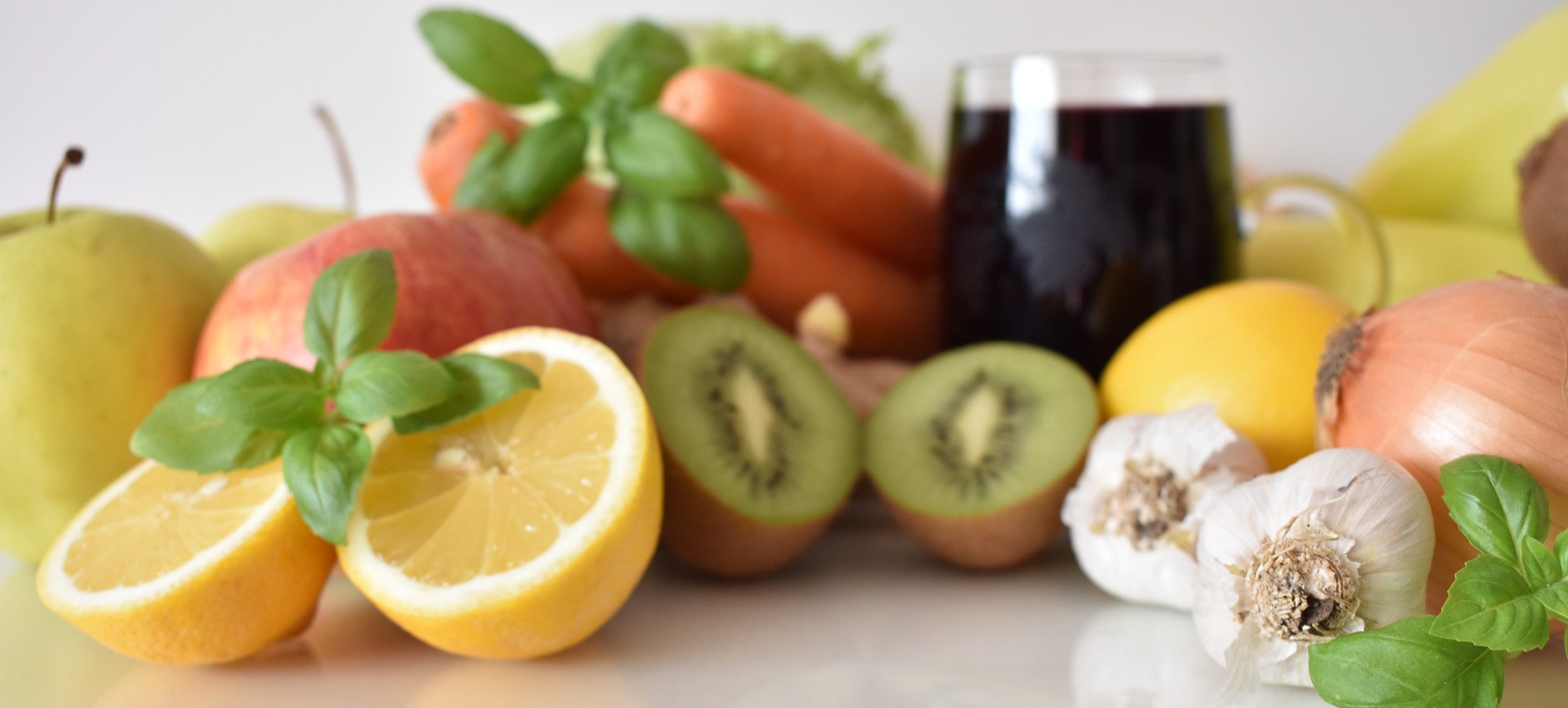 A variety of fruits and vegetables, including apples, kiwi, lemon, carrots, garlic, and an onion, are arranged around a glass of dark juice. The word "HEALTH" is spelled out with wooden letters in the foreground, highlighting the connection between nutrition and mental health.