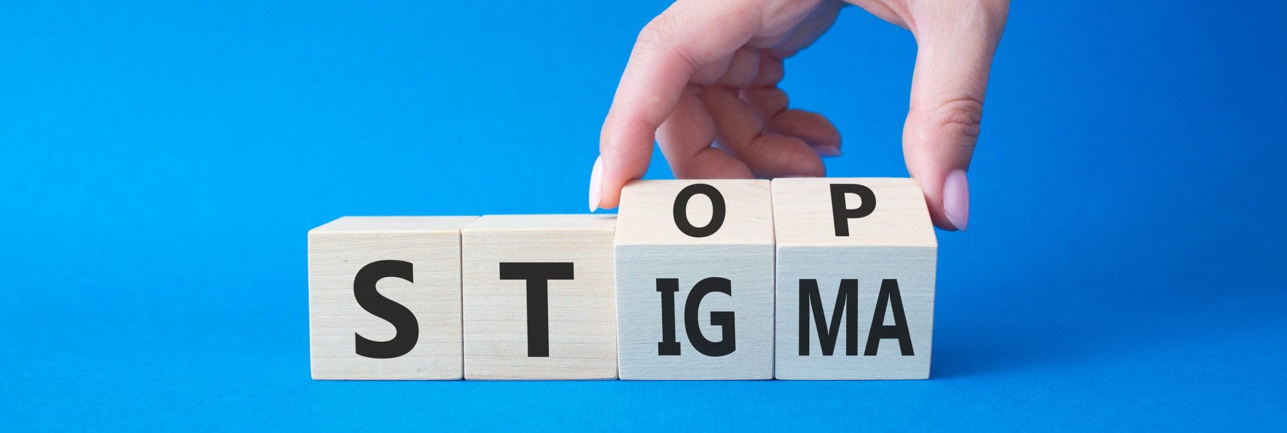 A hand arranges wooden blocks on a blue background, spelling out "STOP STIGMA"—a powerful call to talk about mental health and join in breaking the stigma around it.