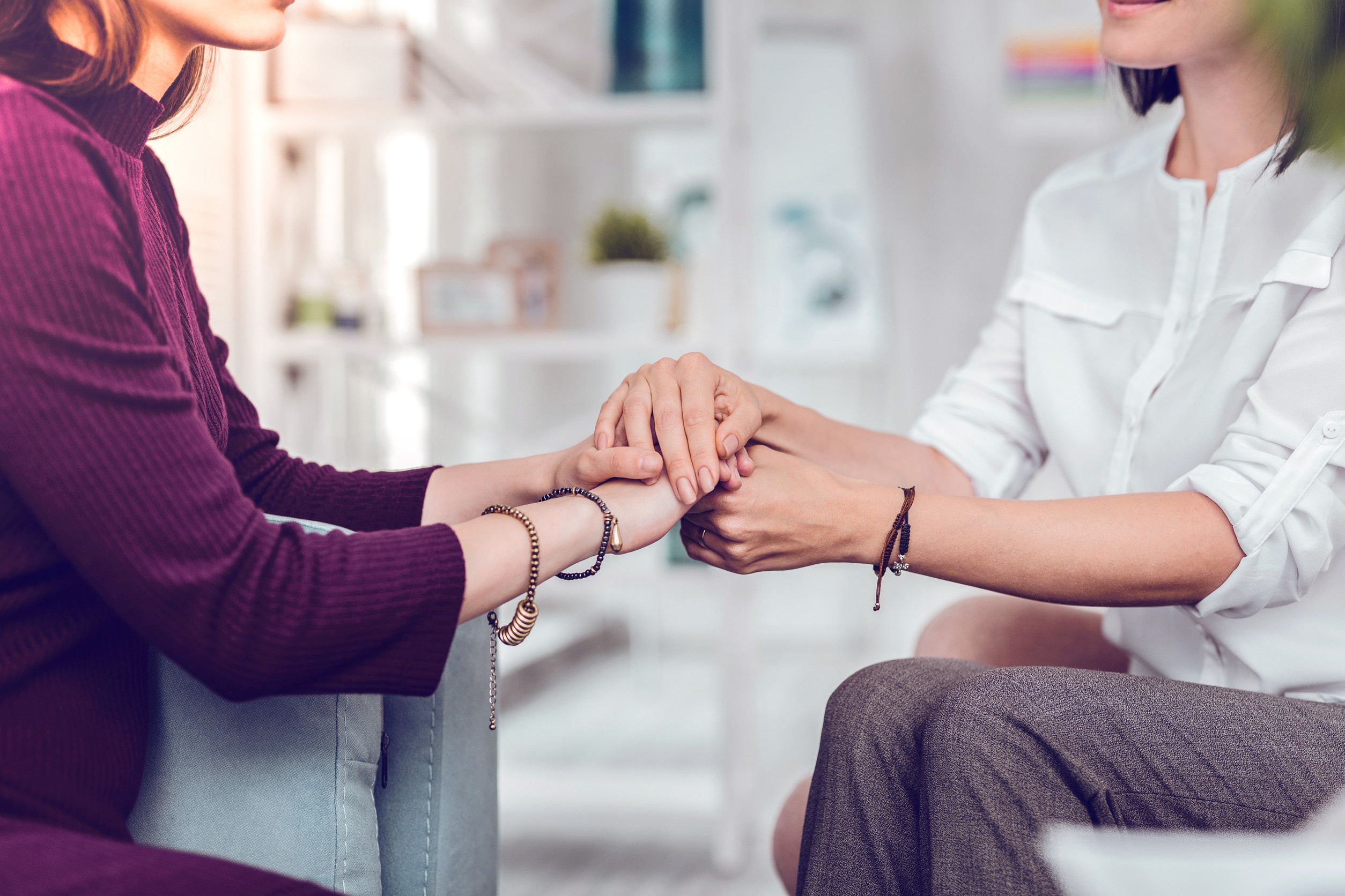 Two women sit close together; one in a purple dress, the other in a white shirt, gently holding hands in a supportive gesture—an intimate moment reflecting compassionate care, perhaps during counseling or psychiatric care, in a calm Minnesota setting.