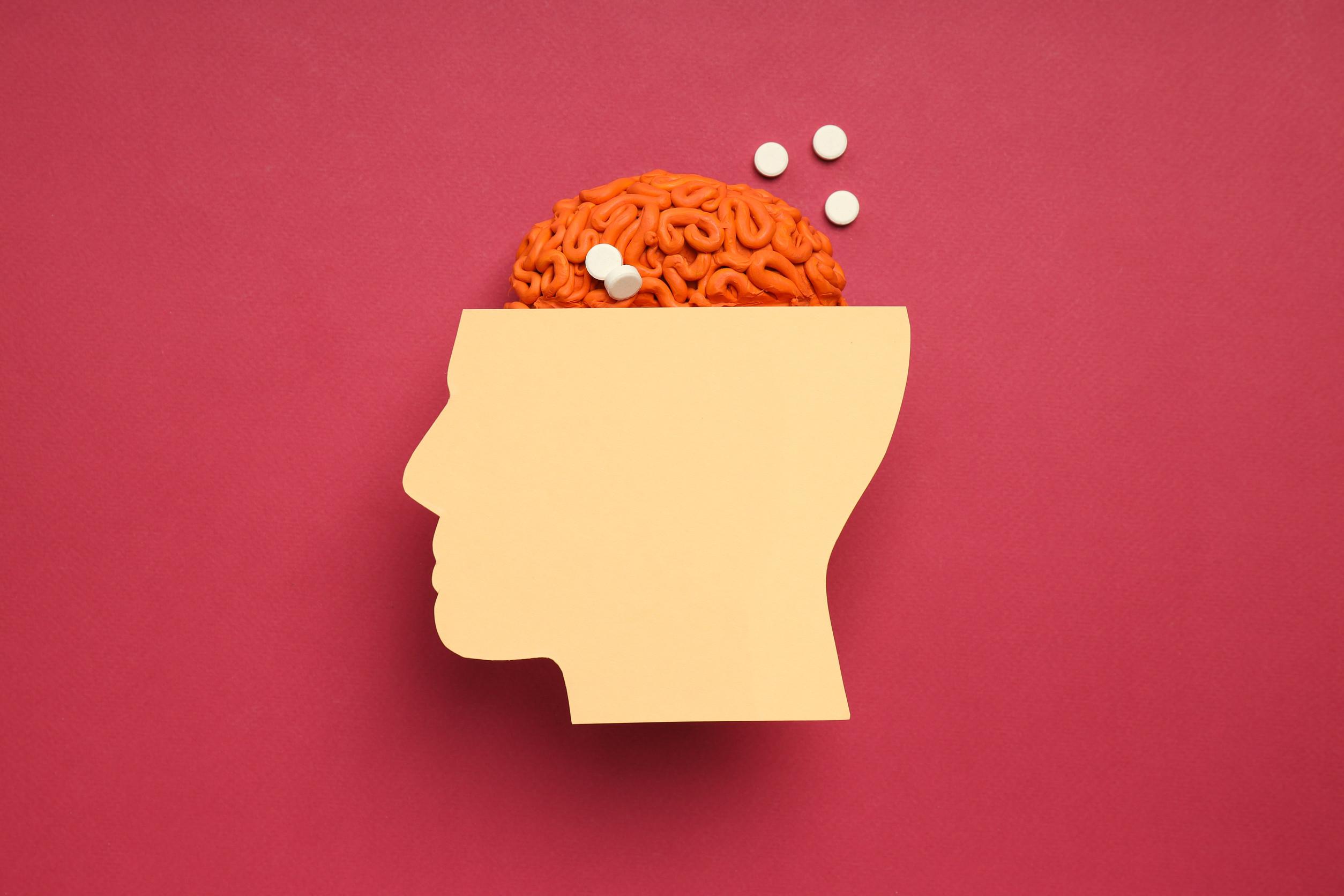 A paper cutout of a human head with an orange brain model inside, topped with several white pills, is set against a solid red background.