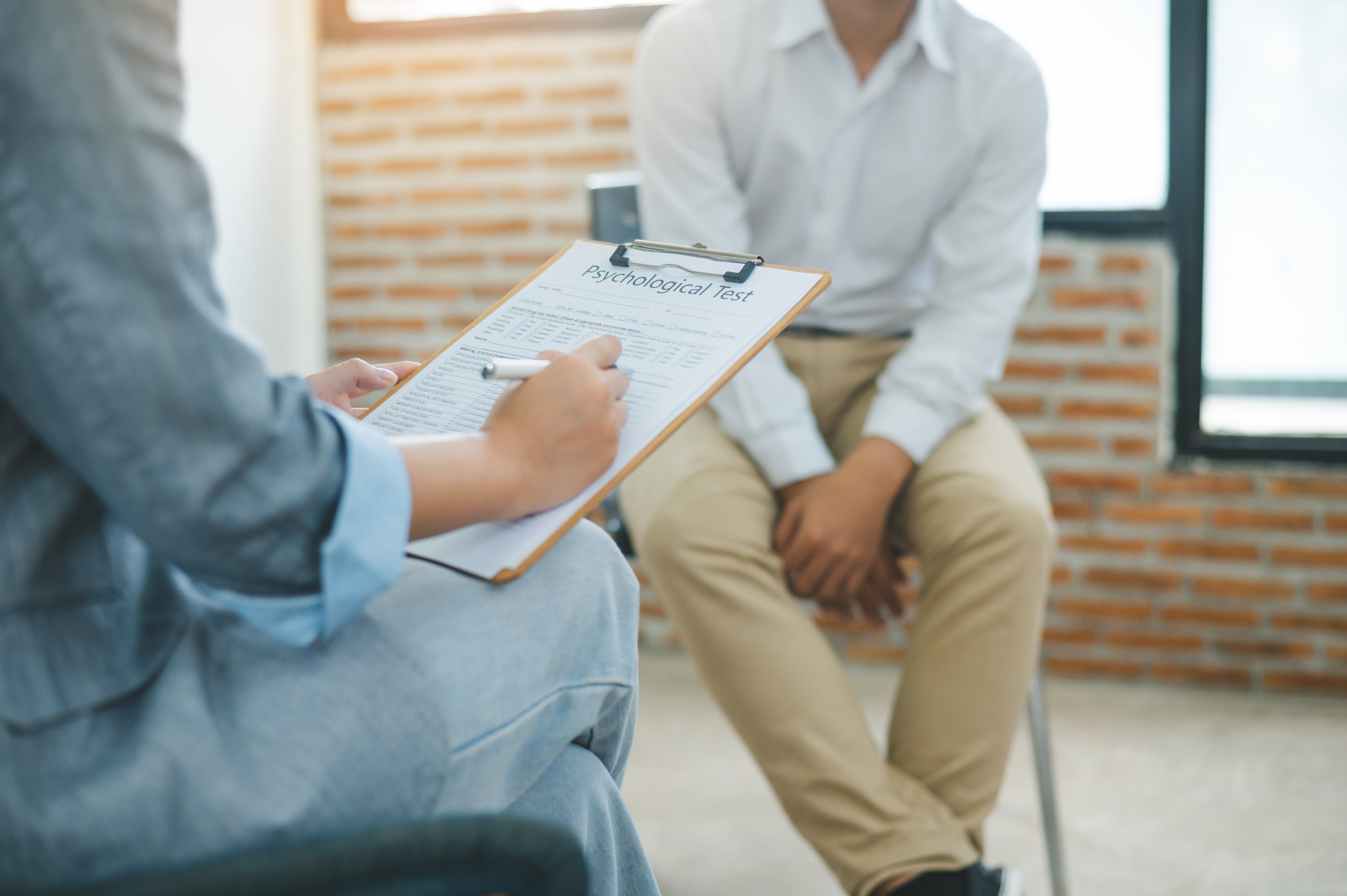 A person holding a clipboard fills out a psychological test form while another prepares for their first psychiatry visit, sitting across with hands clasped in a bright room with a brick wall.