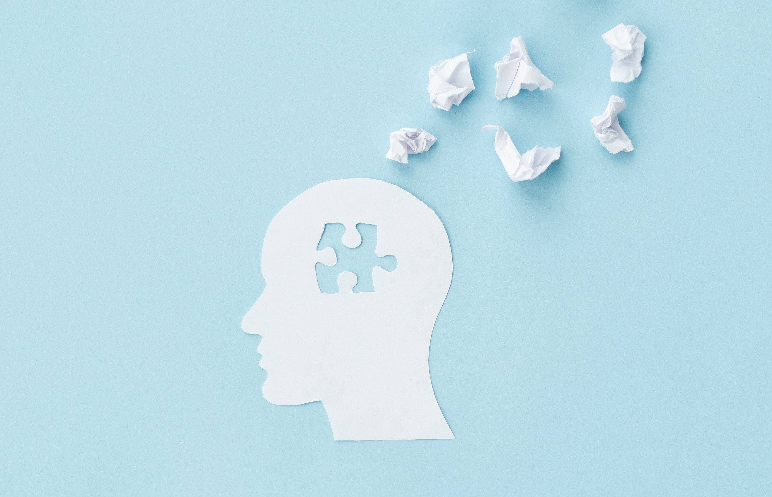 A paper silhouette of a human head with a puzzle piece missing, surrounded by crumpled pieces of paper on a light blue background, symbolizing mental health or problem-solving.
