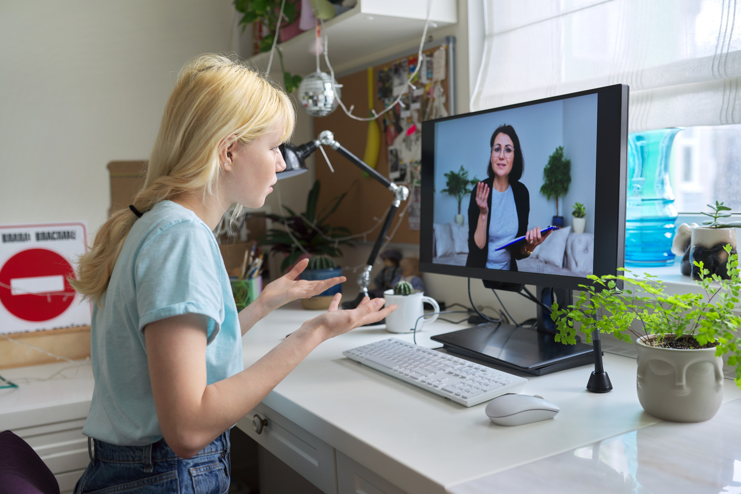 A young woman sits at a desk, gesturing while having a video call with another woman displayed on her computer monitor, using telepsychiatry services in her bright, plant-filled Minnesota home office.