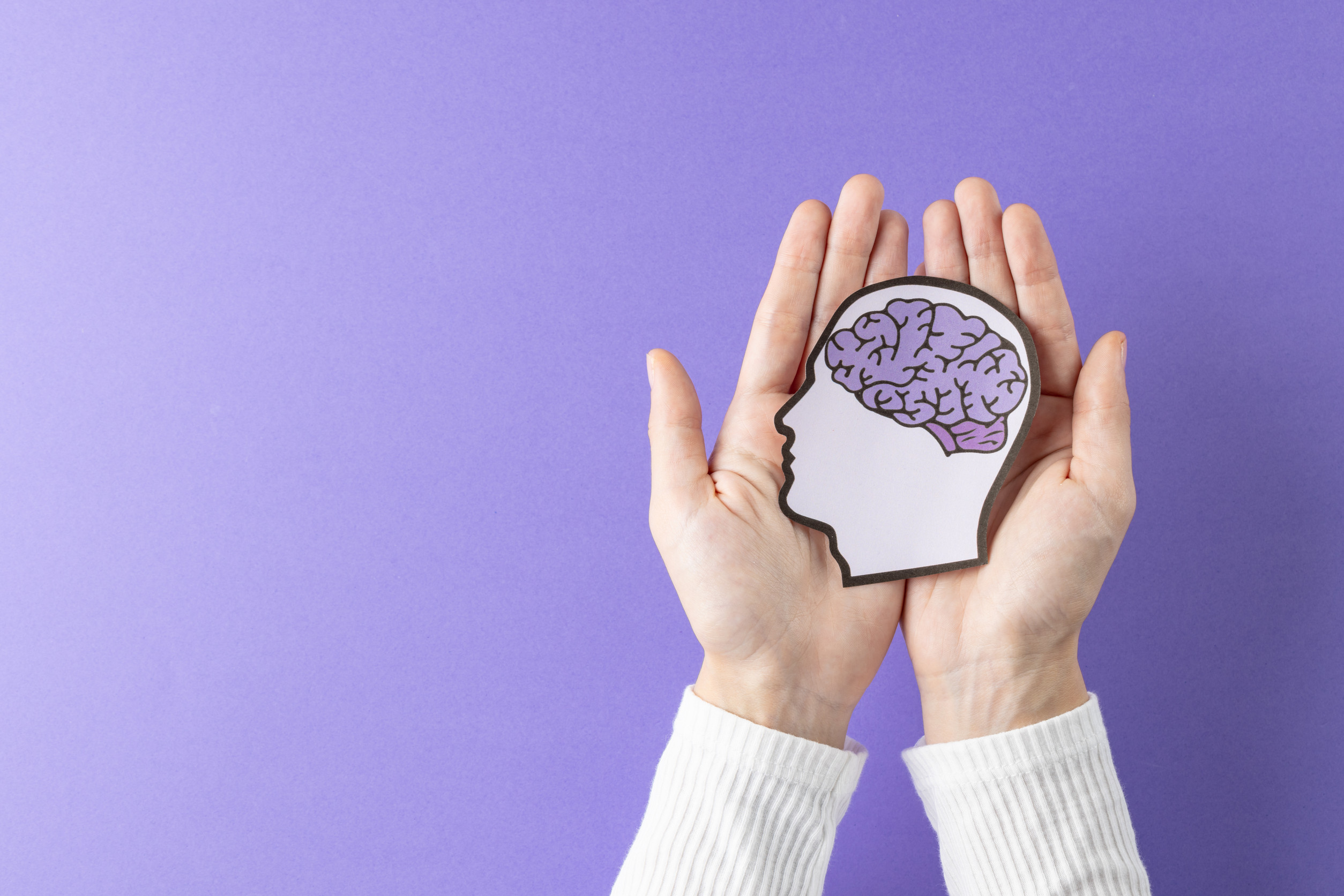 Two hands gently hold a paper cutout of a human head with a brain illustration inside, symbolizing compassion and innovation in mental health care, against a purple background.