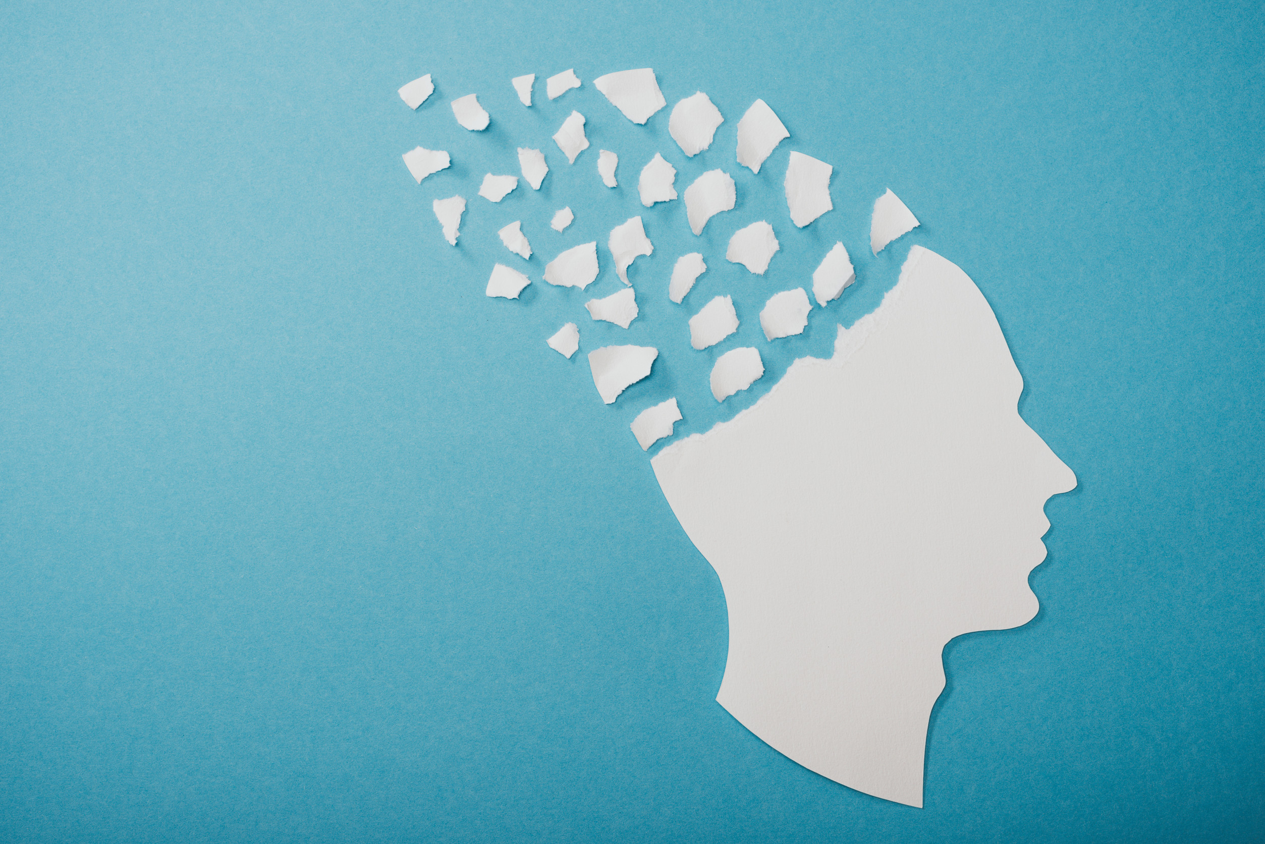 A white paper silhouette of a human head on a blue background, with the top part torn away into scattered pieces, symbolizing memory loss or cognitive decline and reflecting the importance of compassionate care in healing minds.
