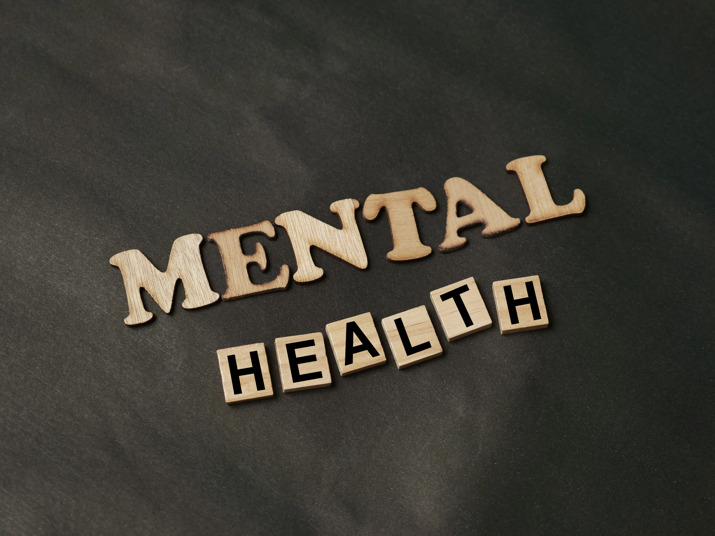 Wooden letters spelling "MENTAL HEALTH" are arranged on a dark gray background, with "MENTAL" in larger wooden letters and "HEALTH" in smaller, block-style letters.