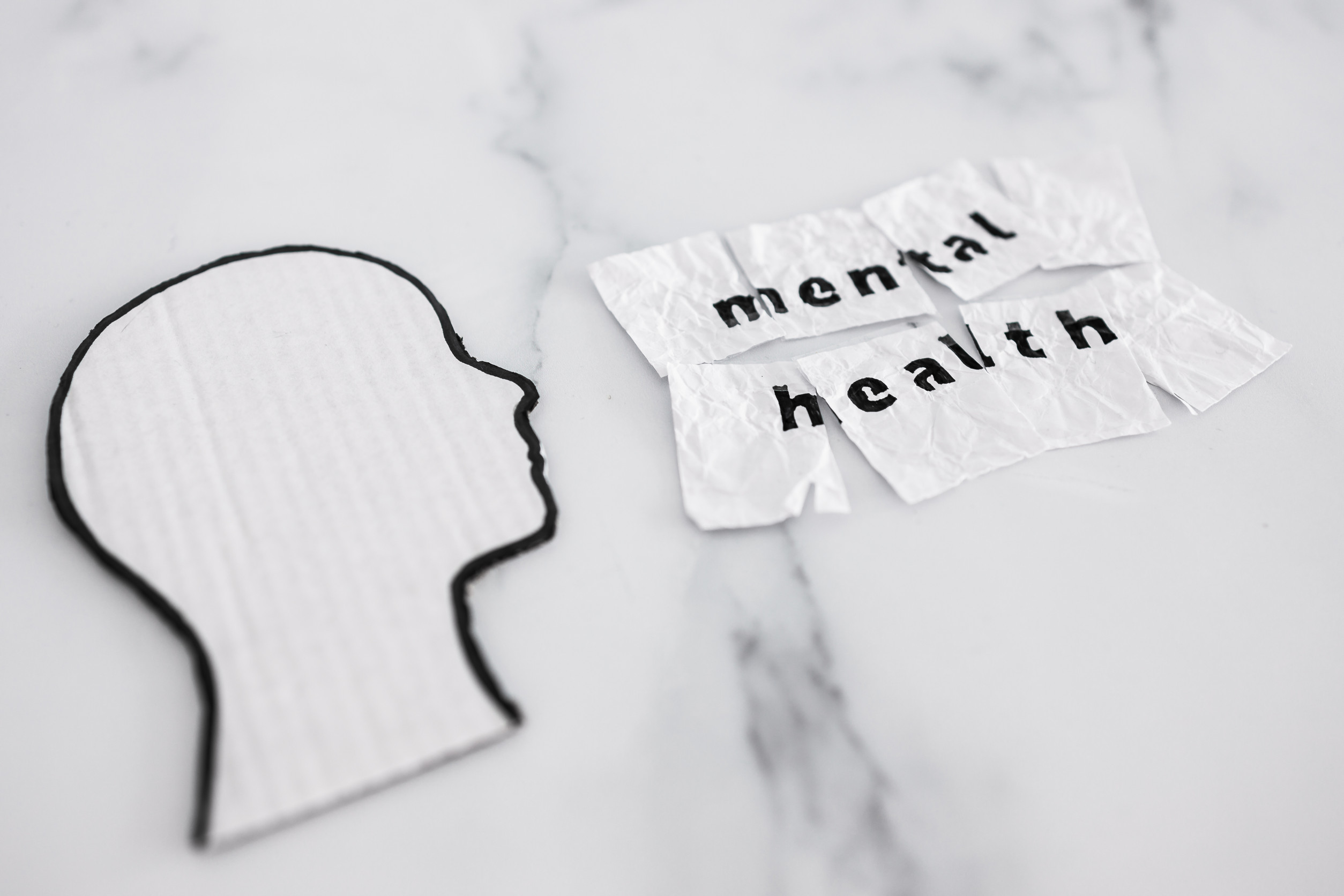 A cutout silhouette of a human head made from white cardboard faces crumpled pieces of paper with the words "mental health" written on them, placed on a white marble surface.