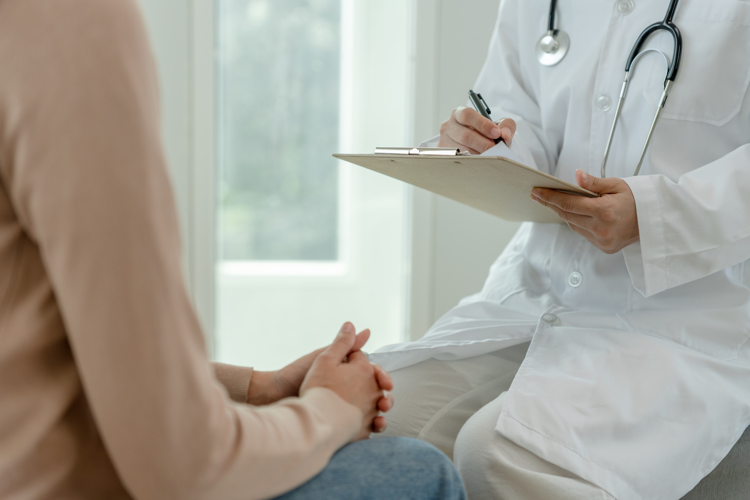 A doctor in a white coat with a stethoscope writes on a clipboard while talking to a seated patient receiving psychiatric disorder care. The setting appears to be a bright medical office in Brainerd.