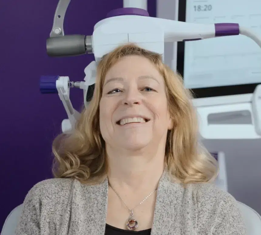 A smiling woman sits in front of a medical device with a gray headpiece positioned near her head, likely for a brain or neurological scan, with purple and white equipment visible in the background.