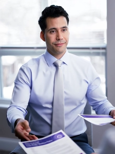 A man in a light blue shirt and tie sits at a desk, holding out a document. He appears to be in an office setting with a window in the background, looking attentive and engaged.