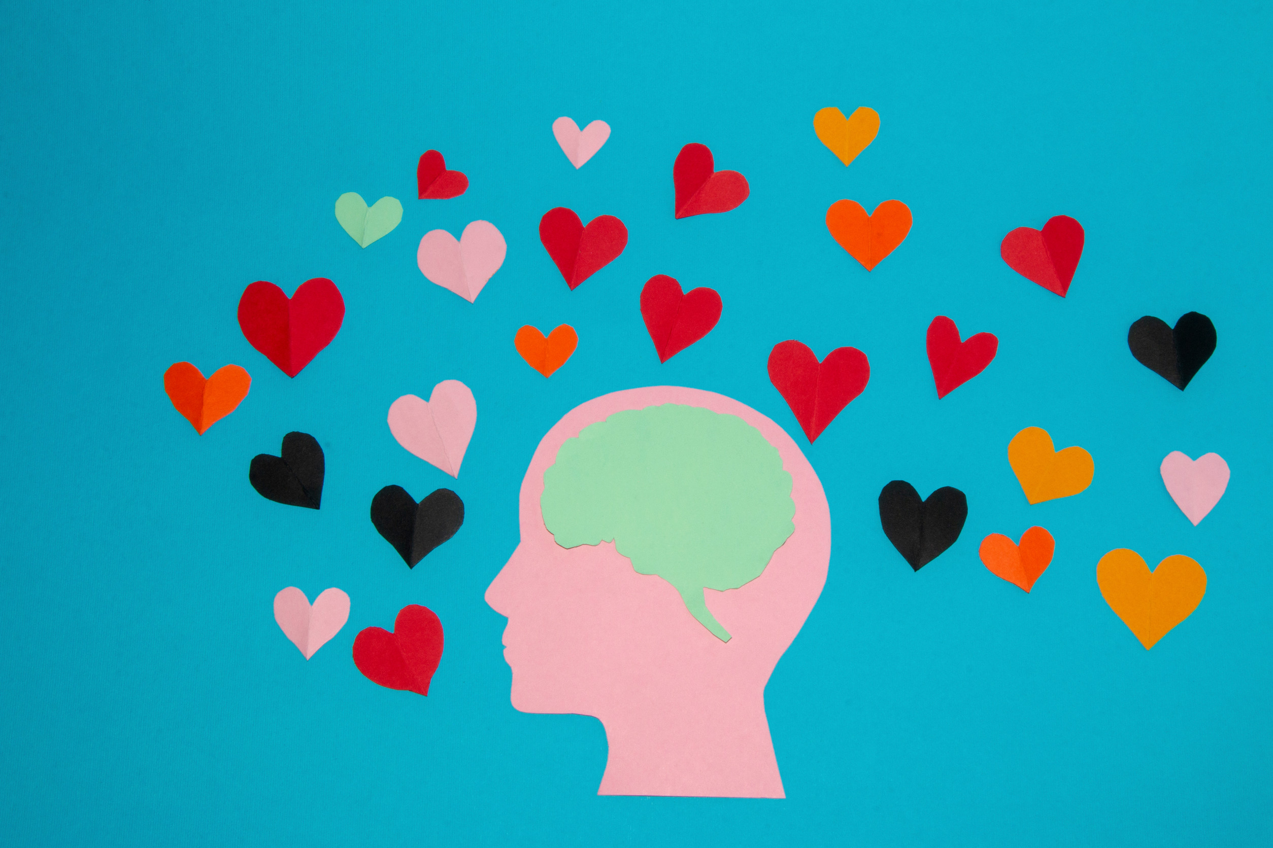 A pink paper silhouette of a head with a green brain is centered on a blue background, symbolizing the mental health journey, and surrounded by colorful paper hearts in red, black, pink, and orange.