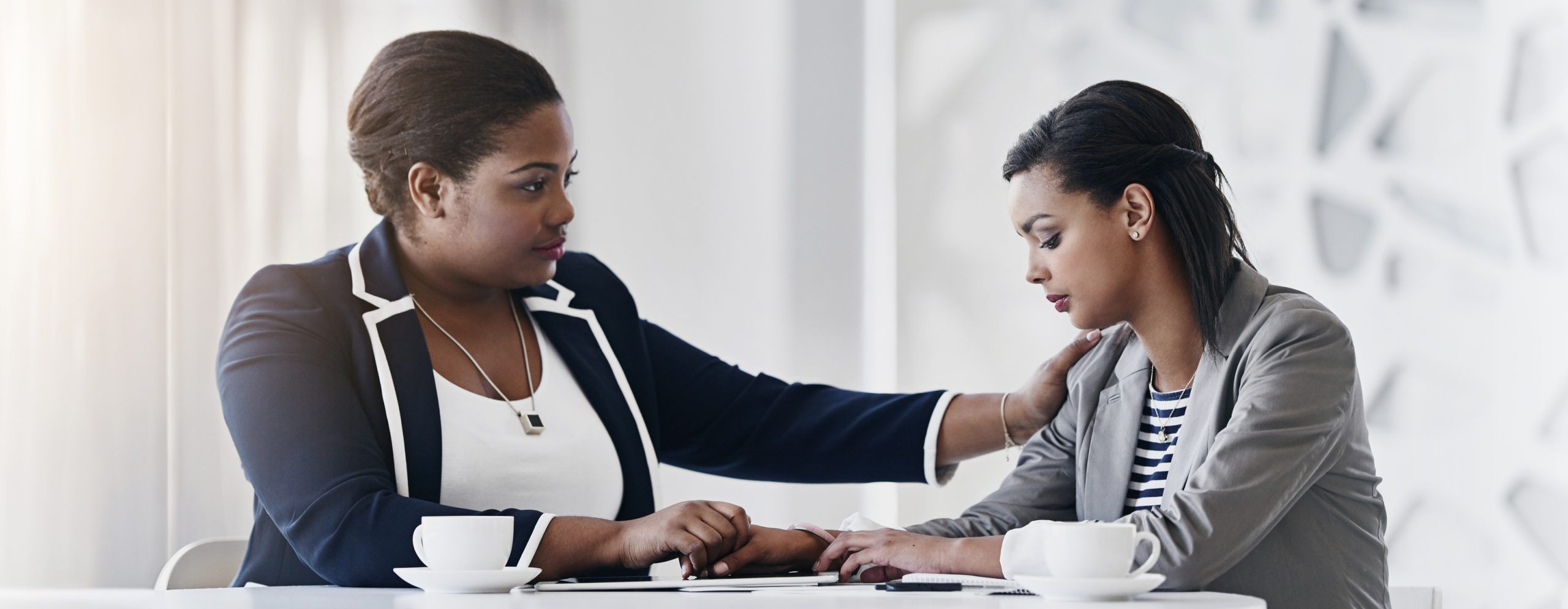 Two women sit at a table with coffee cups; one woman gently comforts the other by placing a hand on her shoulder, while the other looks down with a sad expression.