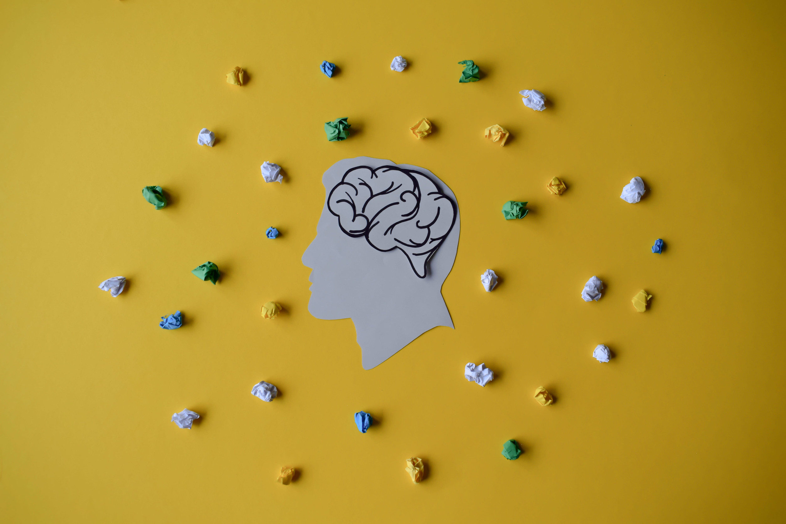 A paper cutout of a human head with a brain illustration is centered on a yellow background, surrounded by small, crumpled balls of paper—symbolizing thoughts—highlighting the journey toward modern anxiety treatment to help you feel better.