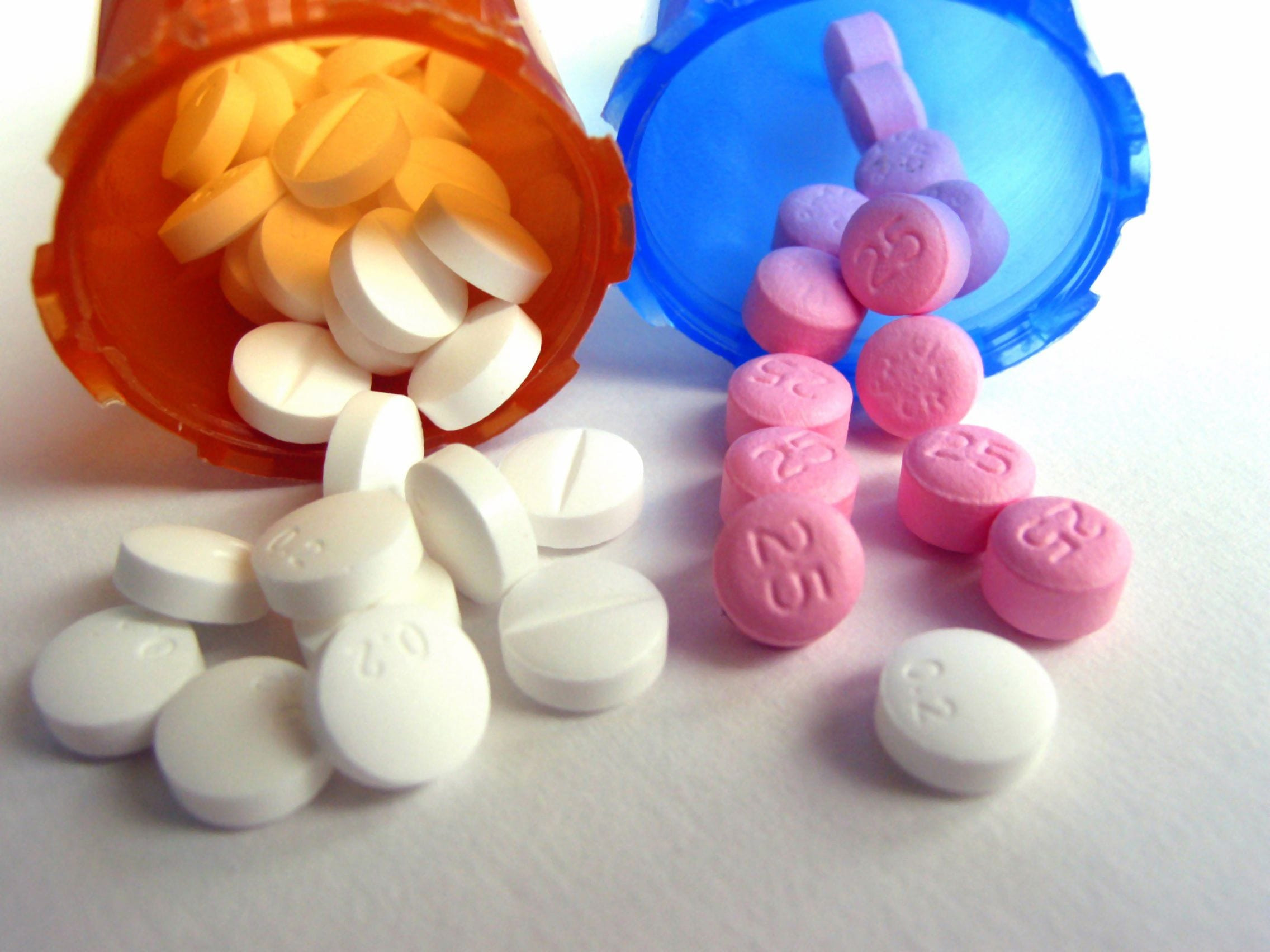 Two prescription pill bottles, one orange and one blue, are tipped over with various round white, pink, and purple tablets spilling out—a visual reference for Medication Management in Minnesota. The pills have numbers and scores imprinted on them.