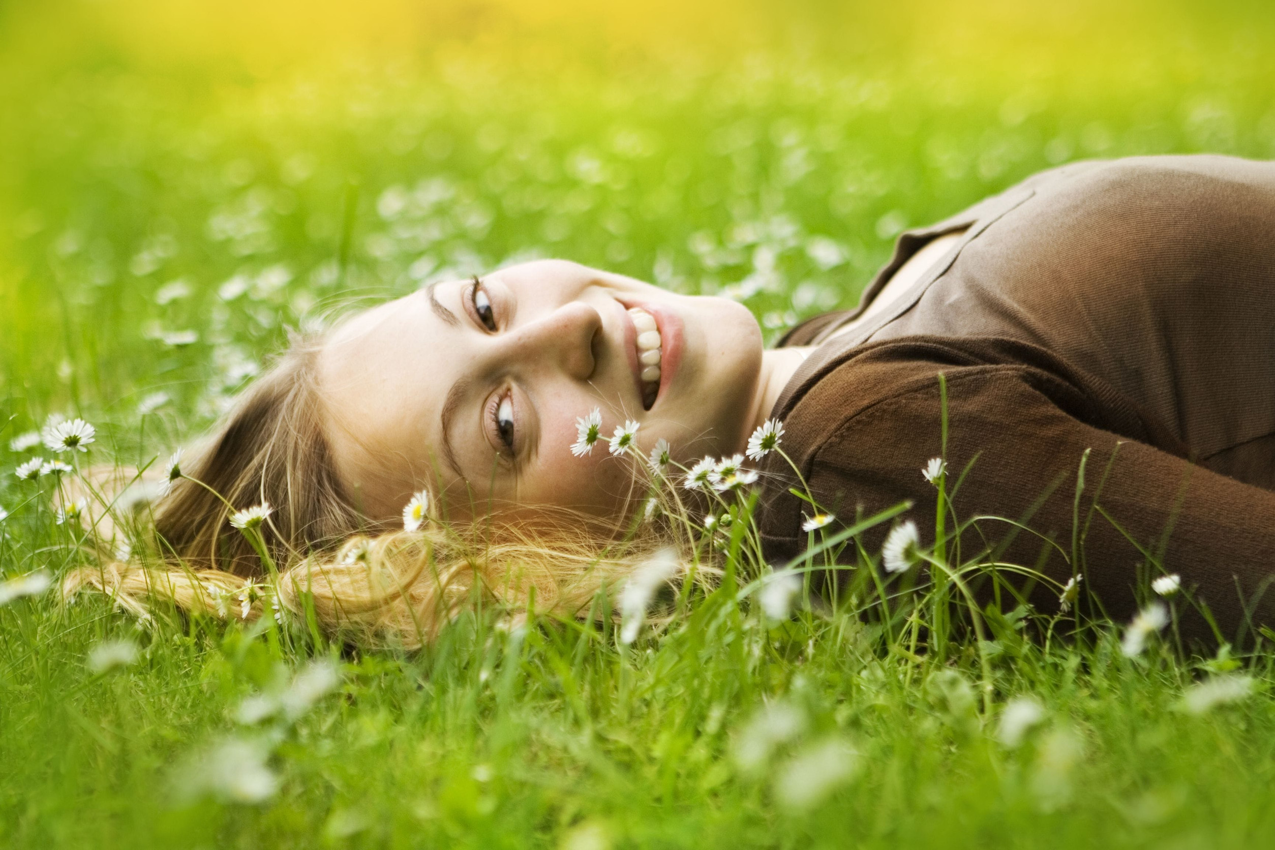 A young woman with blonde hair lies on her side in a grassy field filled with small white flowers, smiling gently at the camera—capturing the warm, sunny hope that comes from long lasting recovery after PTSD treatment.