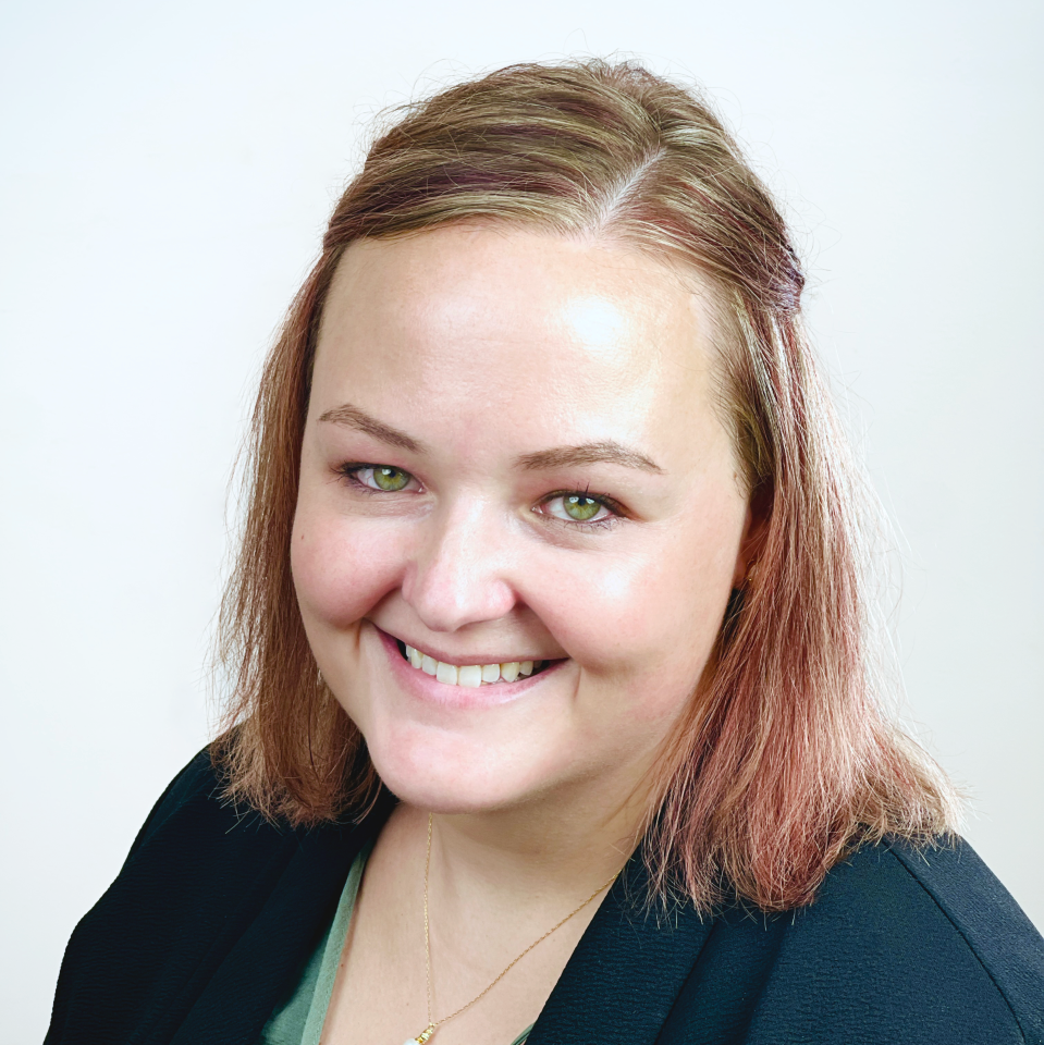 Smiling woman with straight, shoulder-length light brown hair and green eyes, wearing a dark blazer over a green top, posed against a plain light background—ideal for profiles of providers and professionals.