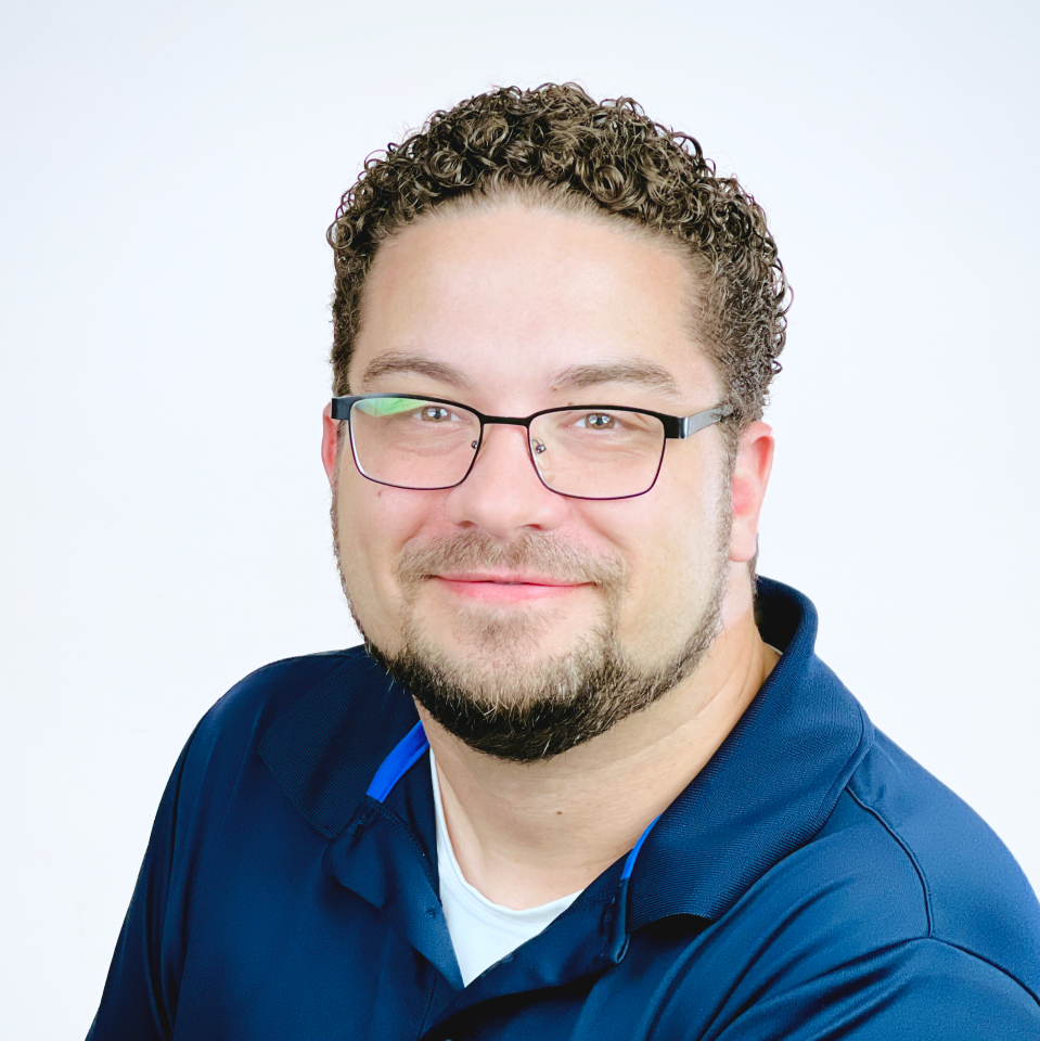 Smiling man with short curly hair, glasses, and a trimmed beard wearing a navy blue collared shirt over a white undershirt, posed against a plain light background—perfect for profiles of trusted providers.