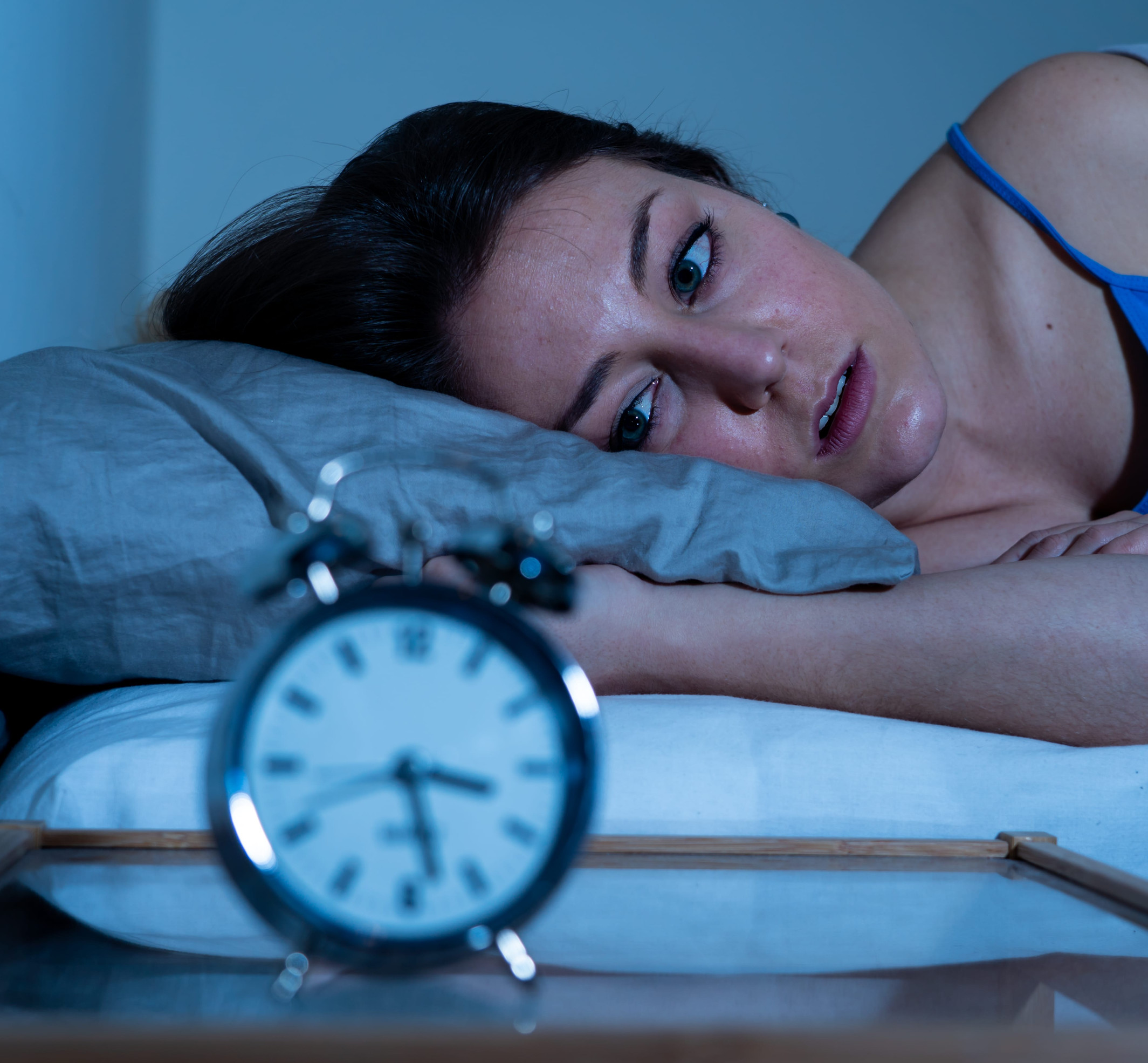 A woman lies in bed with her head on a pillow, looking worriedly at an alarm clock on a bedside table, hinting at sleep problems possibly linked to anxiety. The dimly lit room with blue tones reflects the struggle for rest and mental health concerns.
