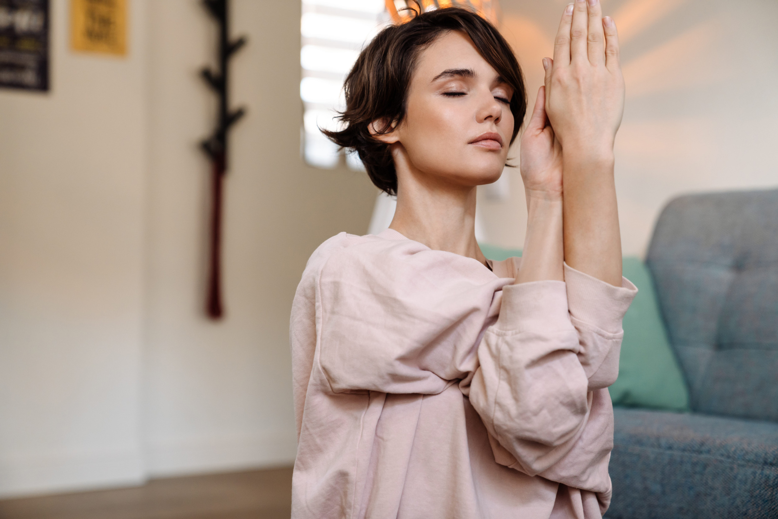 A woman with short brown hair sits indoors, eyes closed, hands together in a supportive yoga or meditation pose. She appears calm and focused, wearing a light pink top—practicing mindfulness for mental health.