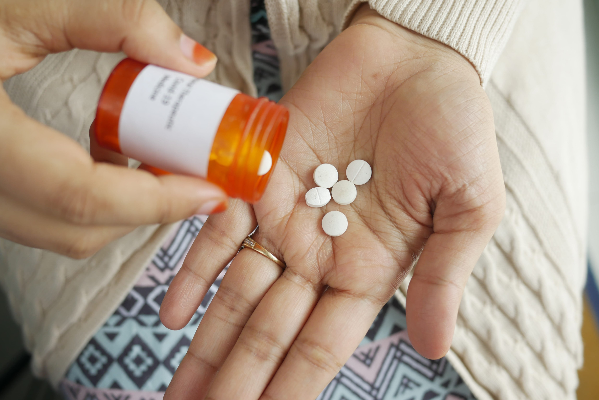 A person pours round white psychiatric medication pills from an orange prescription bottle into their palm, with patterned fabric and a beige sweater visible in the background.