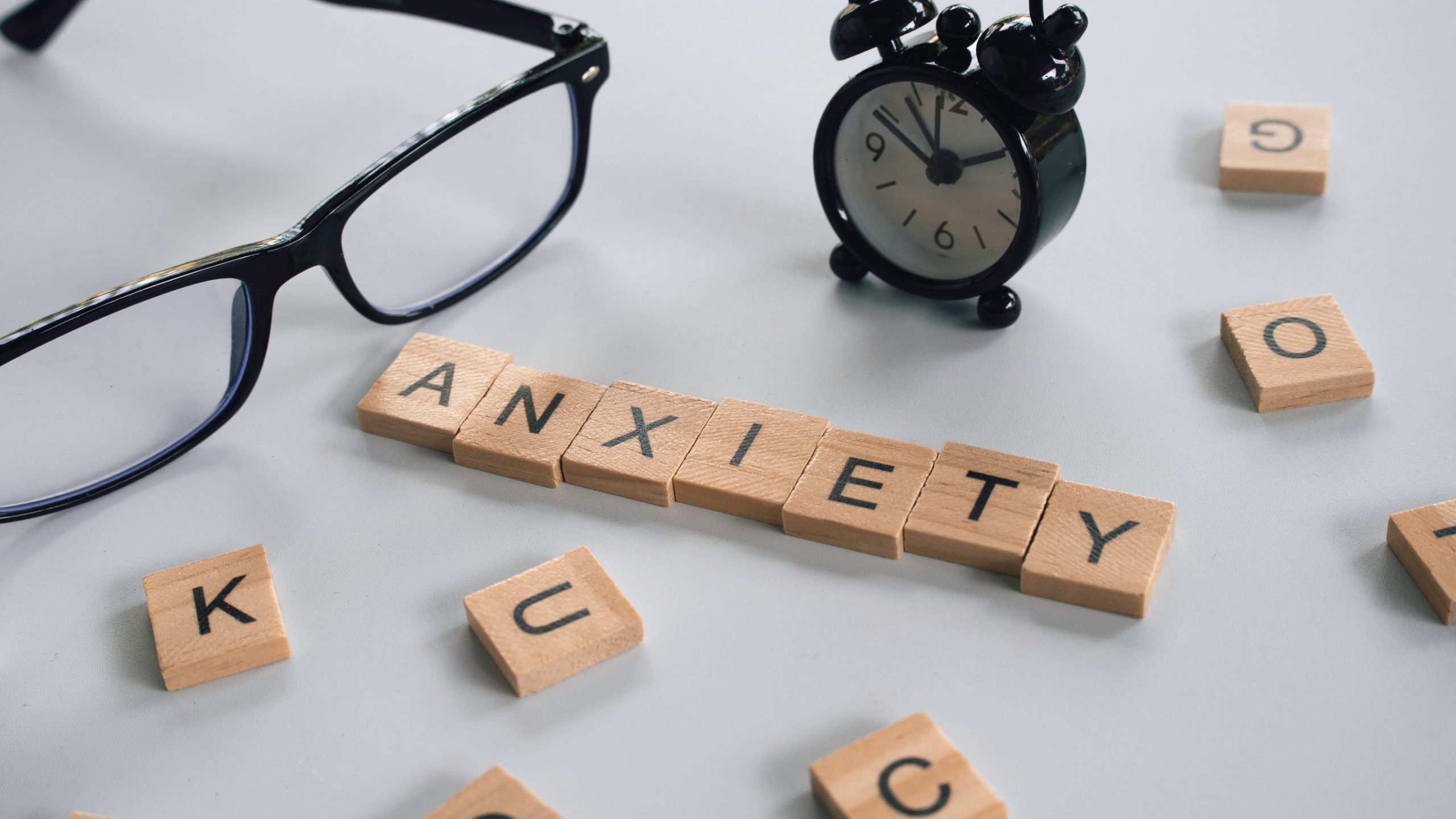 Wooden letter tiles spell "ANXIETY" on a white surface, surrounded by scattered tiles, black eyeglasses, and a small black alarm clock—symbolizing the persistent presence of chronic anxiety in daily life.