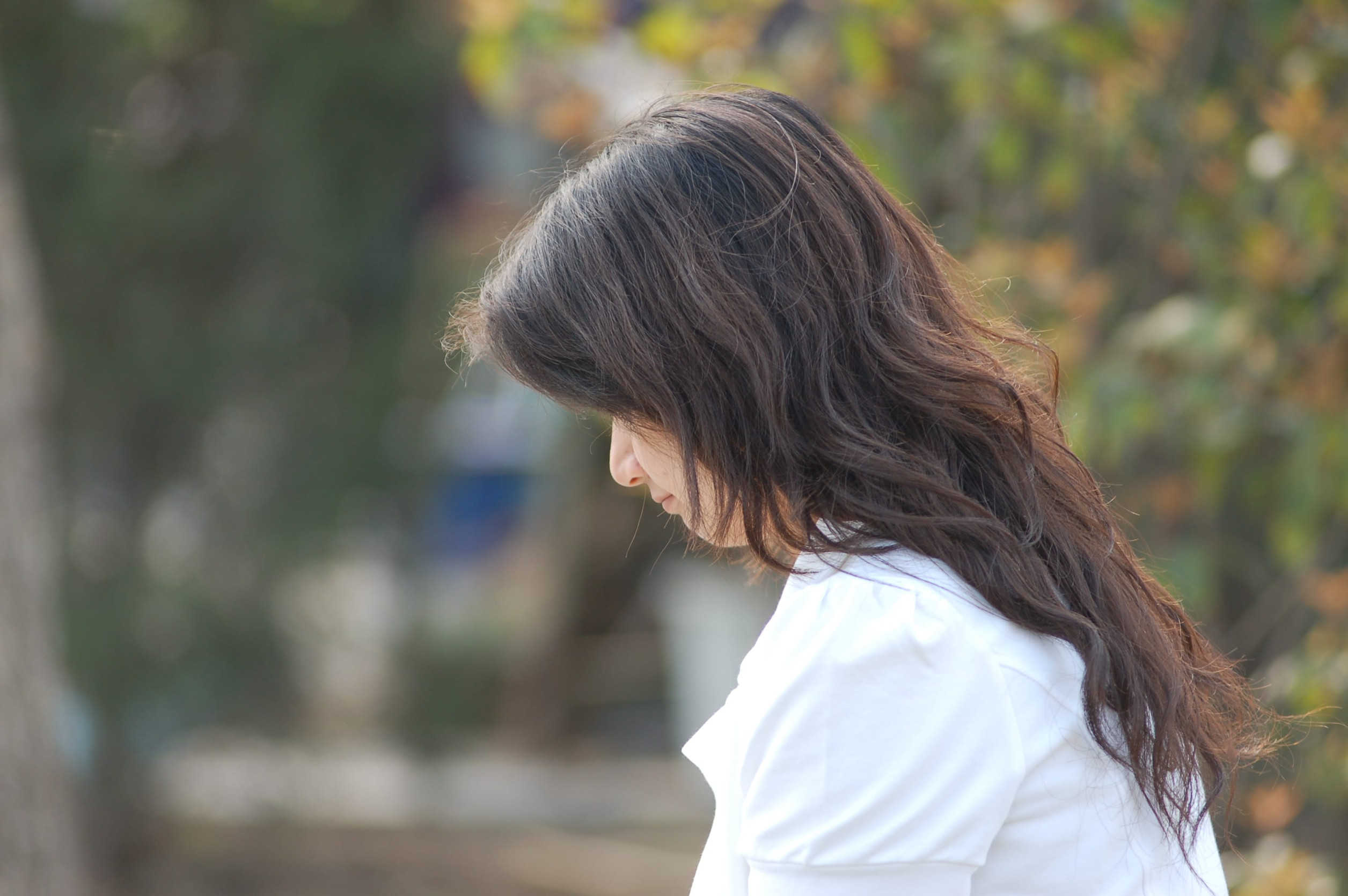 A woman with long brown hair, wearing a white shirt, stands outdoors in profile with her head slightly bowed, surrounded by blurred greenery—a quiet moment of restoring emotional health and finding emotional balance.