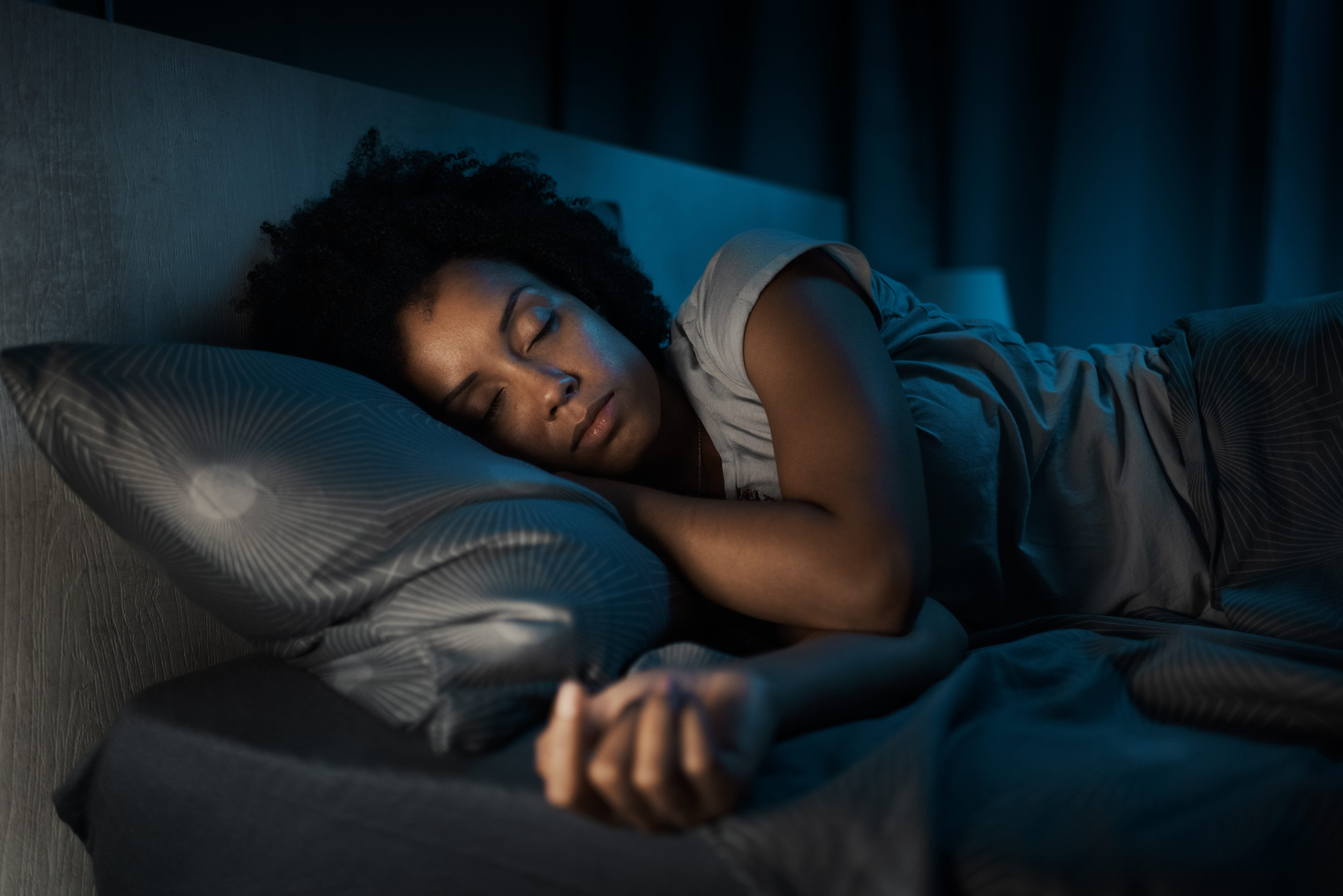 A person with curly hair sleeps peacefully on their side in a dimly lit bedroom, resting their head on a patterned pillow and partially covered with a blanket, highlighting the connection between restful sleep and mental health.