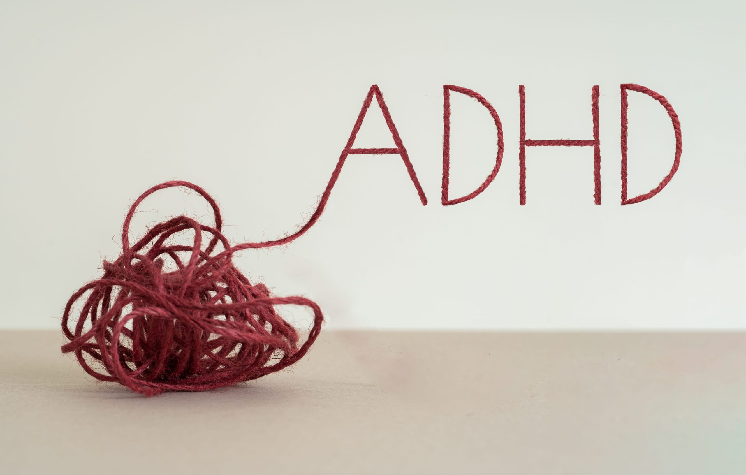 A ball of tangled red yarn extends out to form the letters “ADHD” against a plain background, symbolizing the complexity women often face in receiving ADHD diagnoses.