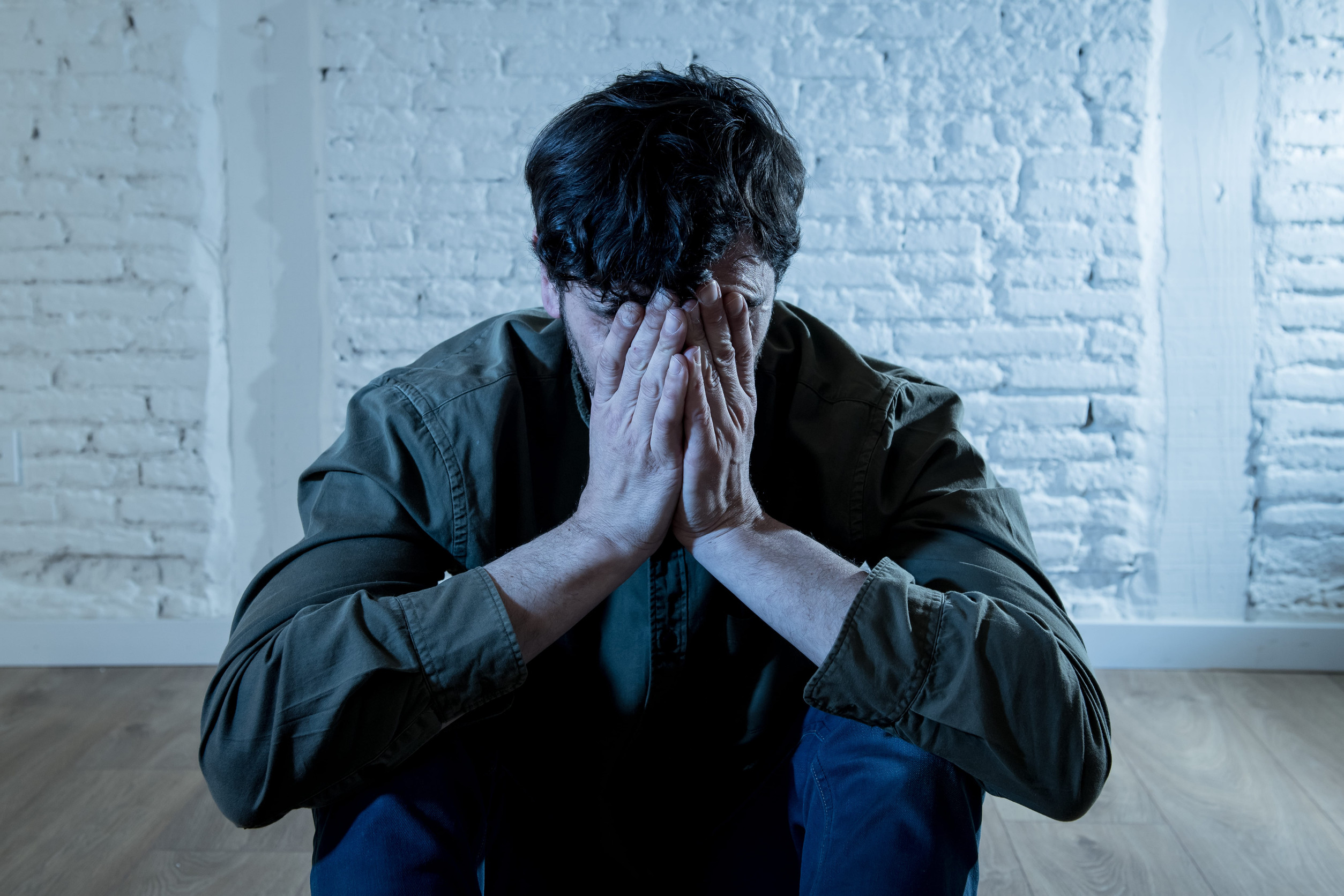 A man sits on the floor against a white brick wall, leaning forward with his elbows on his knees and his hands covering his face, appearing distressed or upset—a posture that may reflect hidden signs of depression.