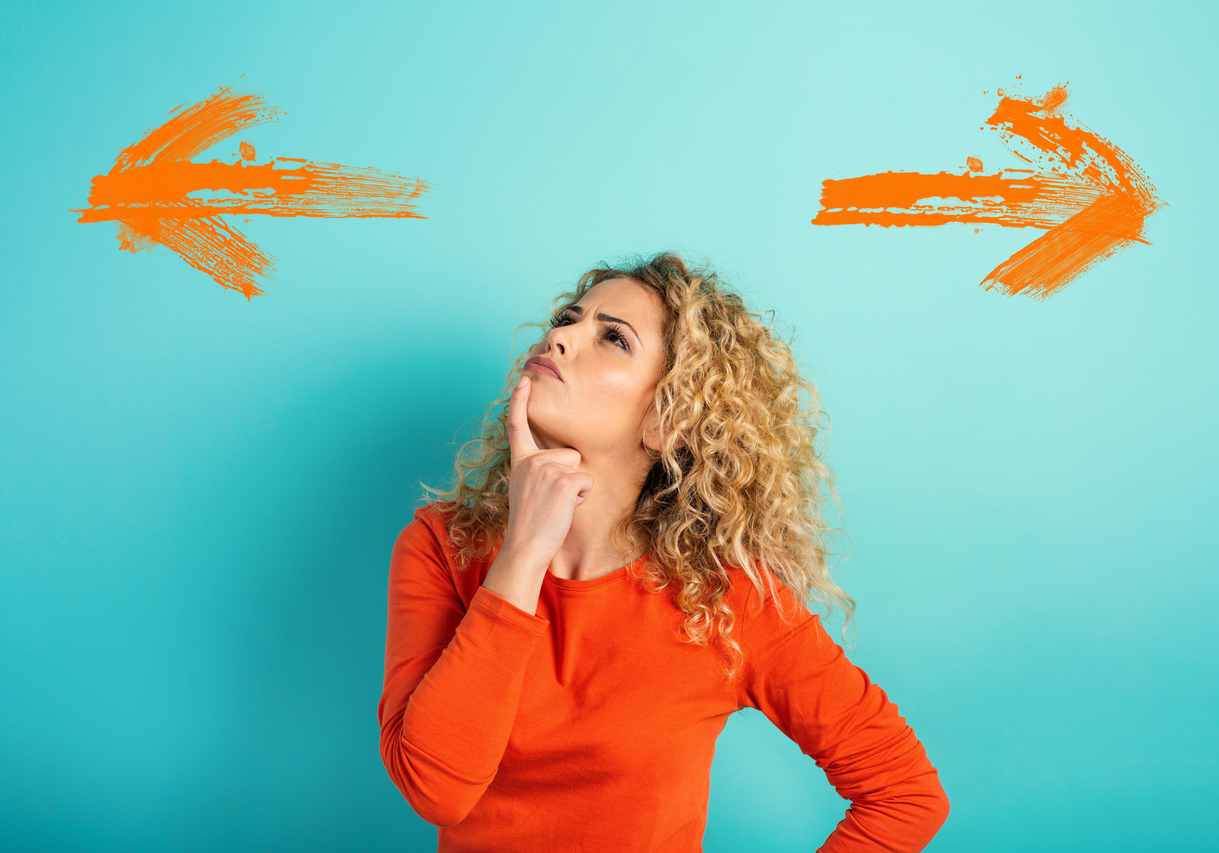 A woman in an orange shirt stands against a blue background, looking thoughtful, with her hand on her chin. Orange arrows point left and right on either side of her, suggesting a decision or choice—perhaps reflecting the process of seeking support for mental health by choosing between seeing a psychiatrist or therapist.