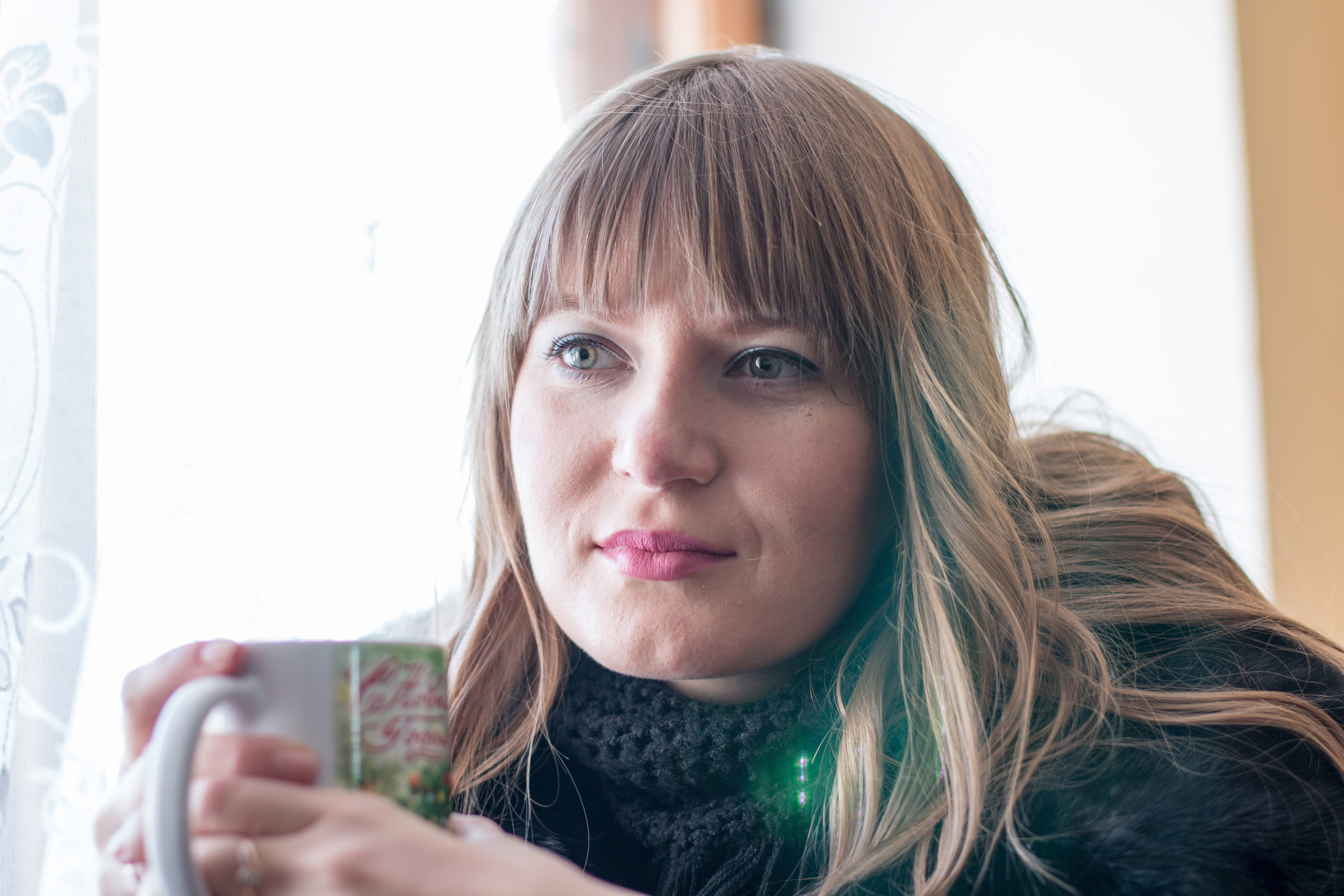 A woman with long light brown hair and bangs sits by a window, holding a mug and looking thoughtfully into the distance. She wears a black scarf and a dark top, appearing to find calm amidst life transitions.