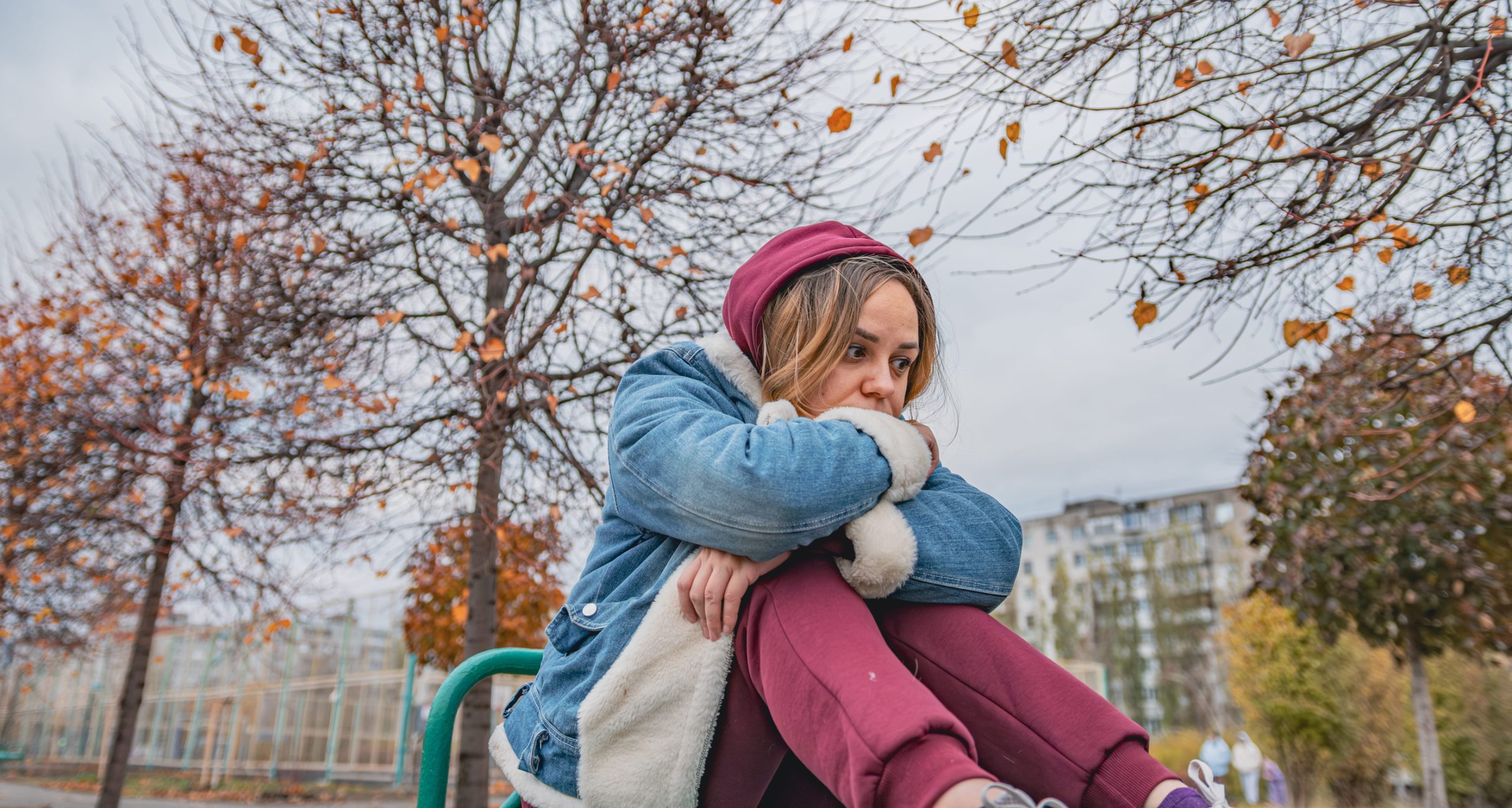 A woman in a denim jacket and maroon hoodie sits outdoors on a bench, looking thoughtful—perhaps reflecting on fall mental health—while leafless trees and apartment buildings frame the cloudy day.