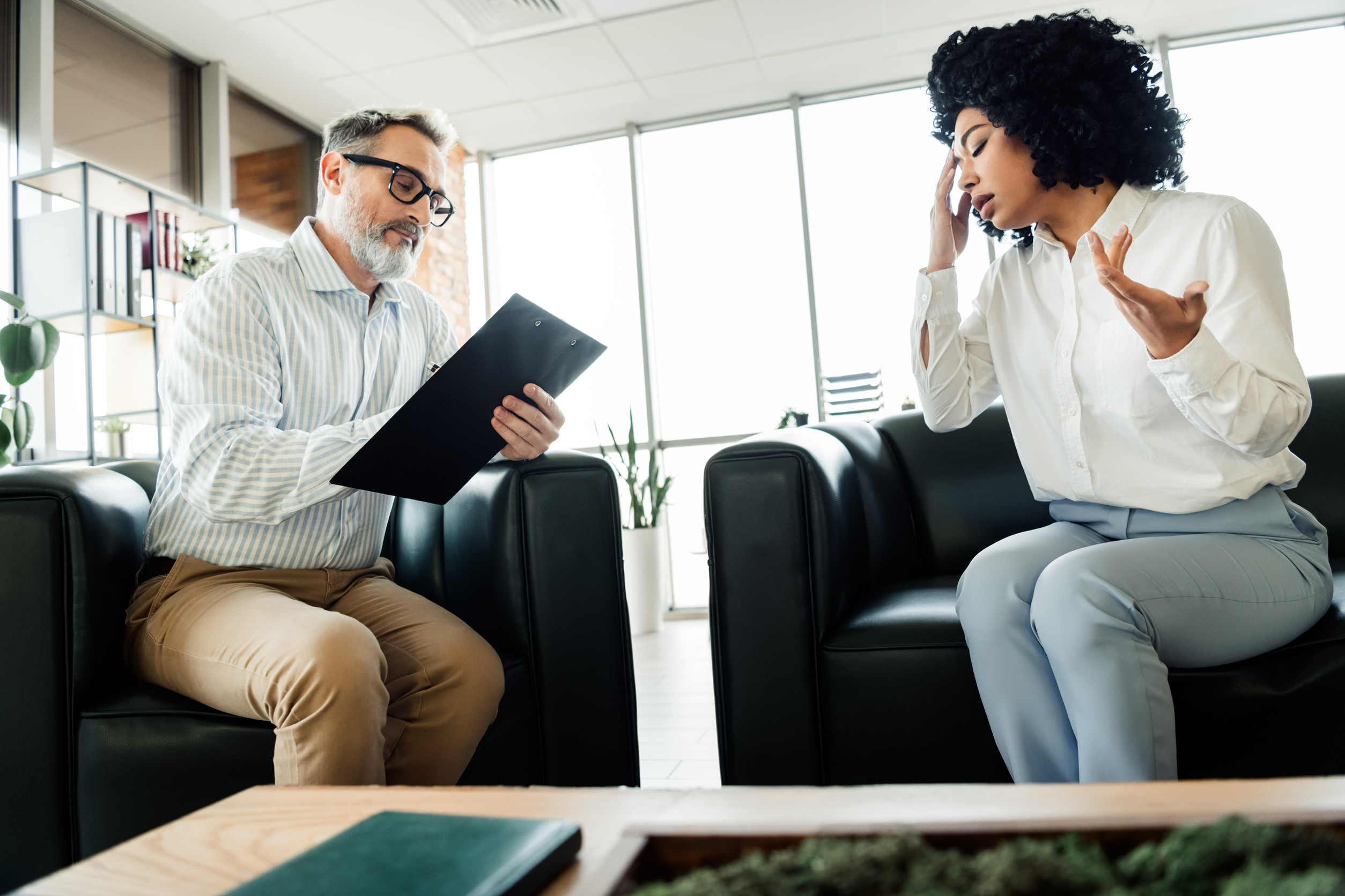 A man holding a clipboard listens attentively to a woman who appears frustrated or upset during her first psychiatric evaluation in a modern office or therapy setting with large windows and plants.