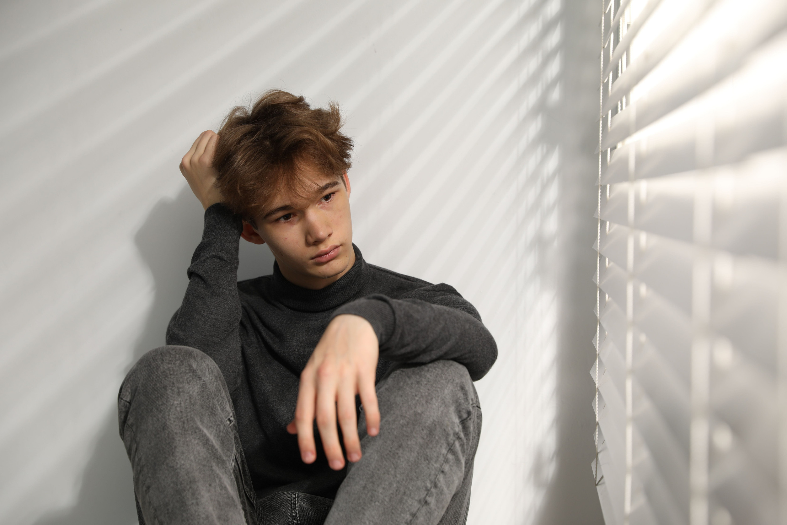 A young person with light brown hair sits against a wall in soft sunlight, casting shadows from window blinds. Wearing a dark long-sleeve shirt and gray jeans, they look thoughtful with one hand in their hair—a quiet moment that reflects the sense of back-to-school anxiety many students experience each year.